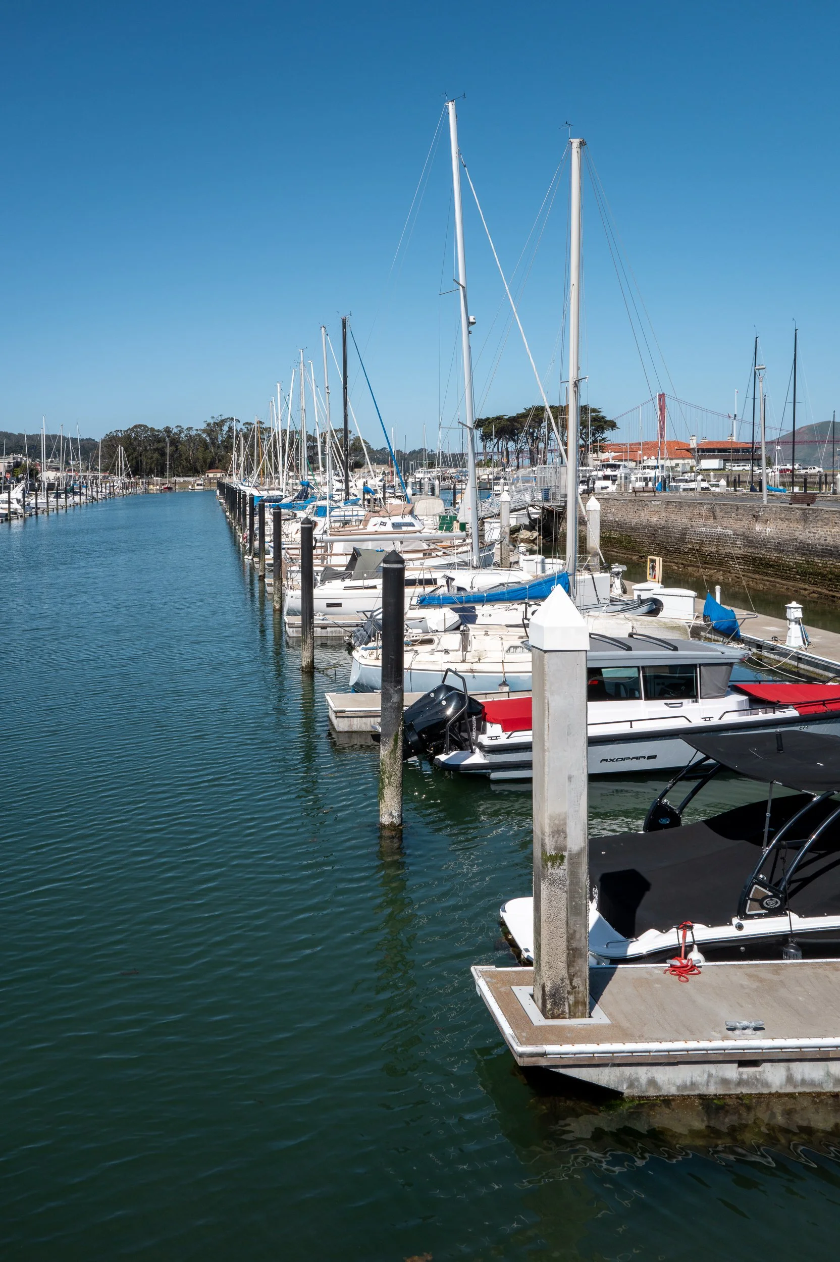 Havre de bateaux avec plusieurs voiliers et bateaux à moteur amarrés, avec un quai en béton et une mer calme sous un ciel bleu clair.