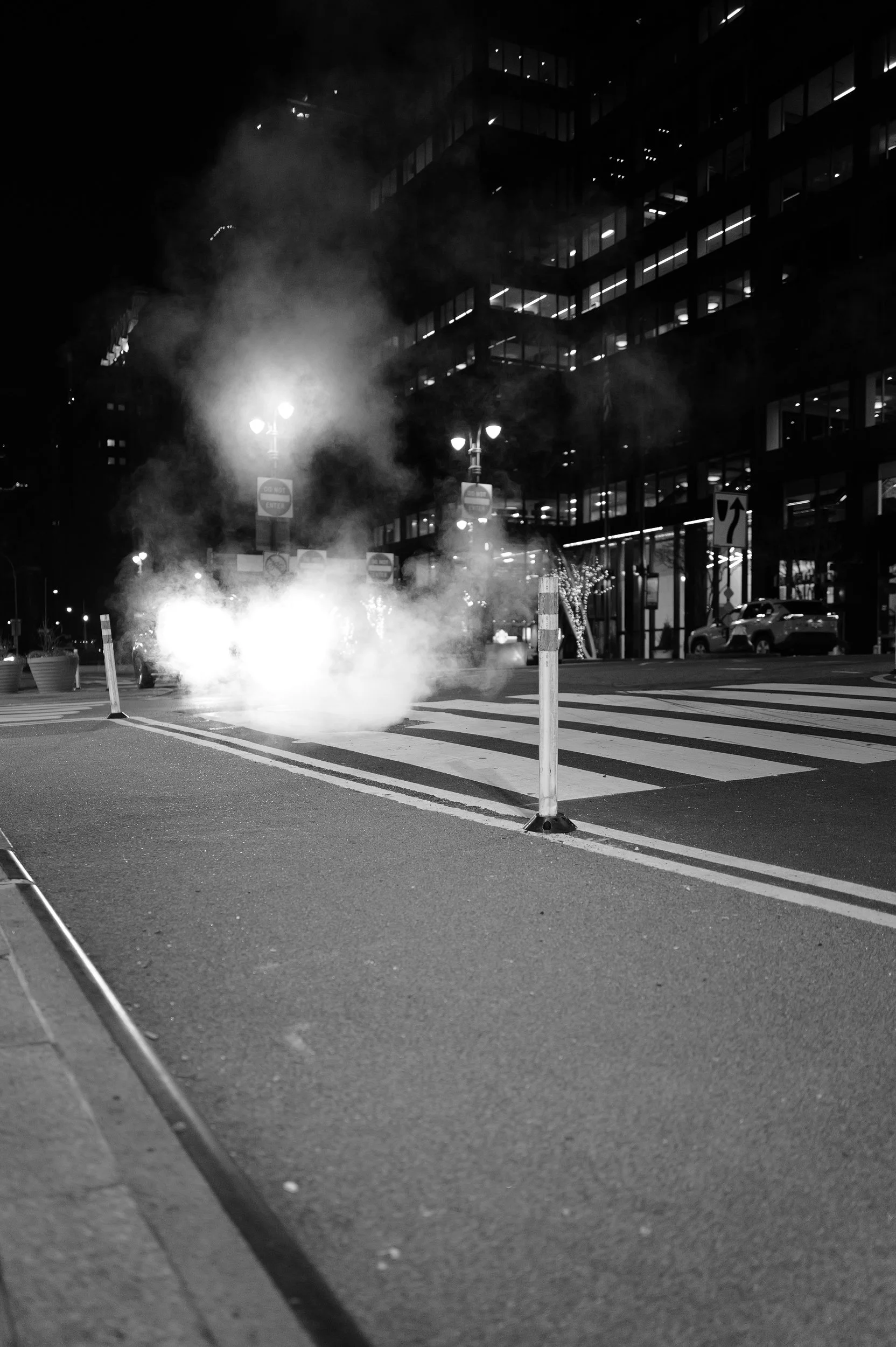 Une rue urbaine la nuit, avec un panneau de contrôle de circulation entouré de fumée ou de vapeur, et des barrières de chantier.