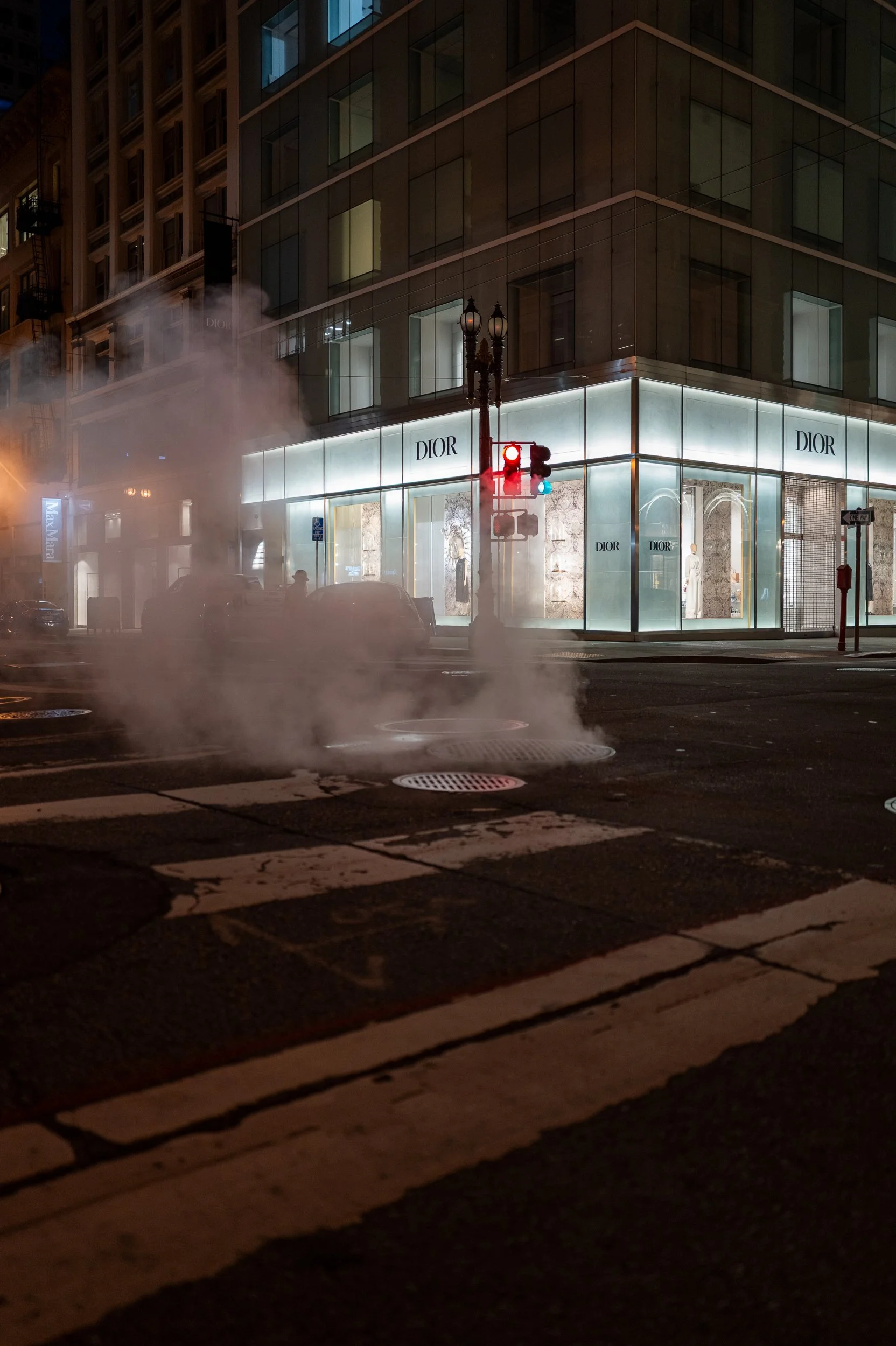 Une rue la nuit avec une boutique Dior illuminée, du brouillard ou de la vapeur s'échappant du sol, un feu de circulation rouge et des bâtiments modernes en arrière-plan.