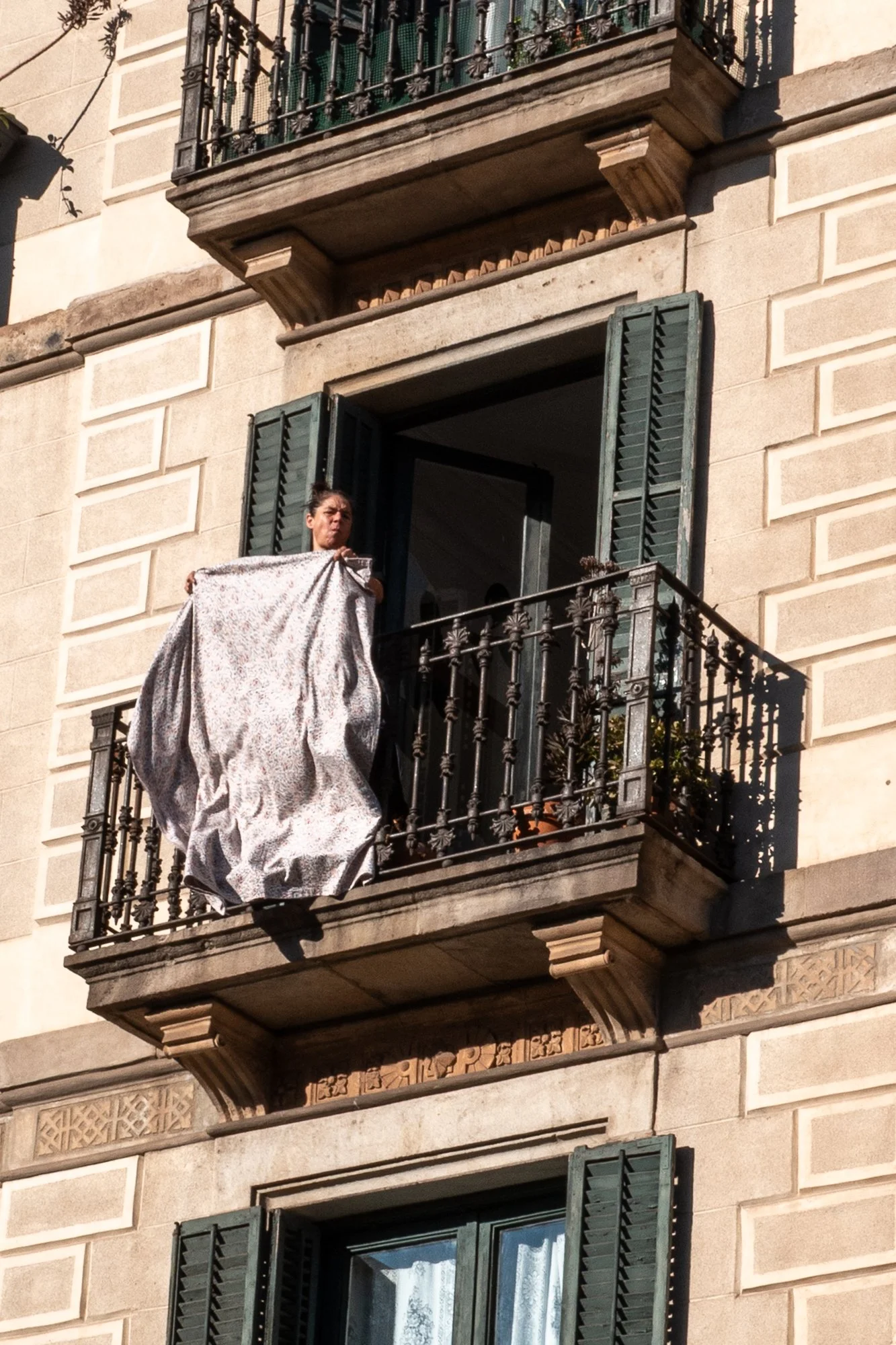 Femme tenant un drap ou une couverture sur un balcon d'un immeuble en pierre avec des volets verts.