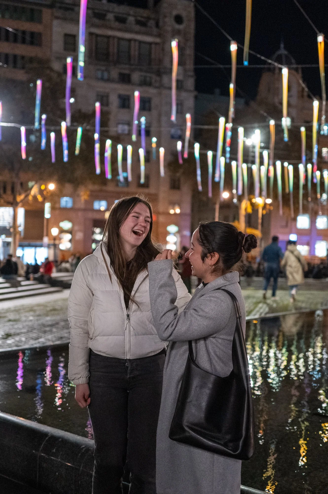 Deux femmes riant et parlant dans une place urbaine illuminée la nuit, avec des décorations colorées suspendues au-dessus d'elles et un reflet dans l'eau derrière elles.