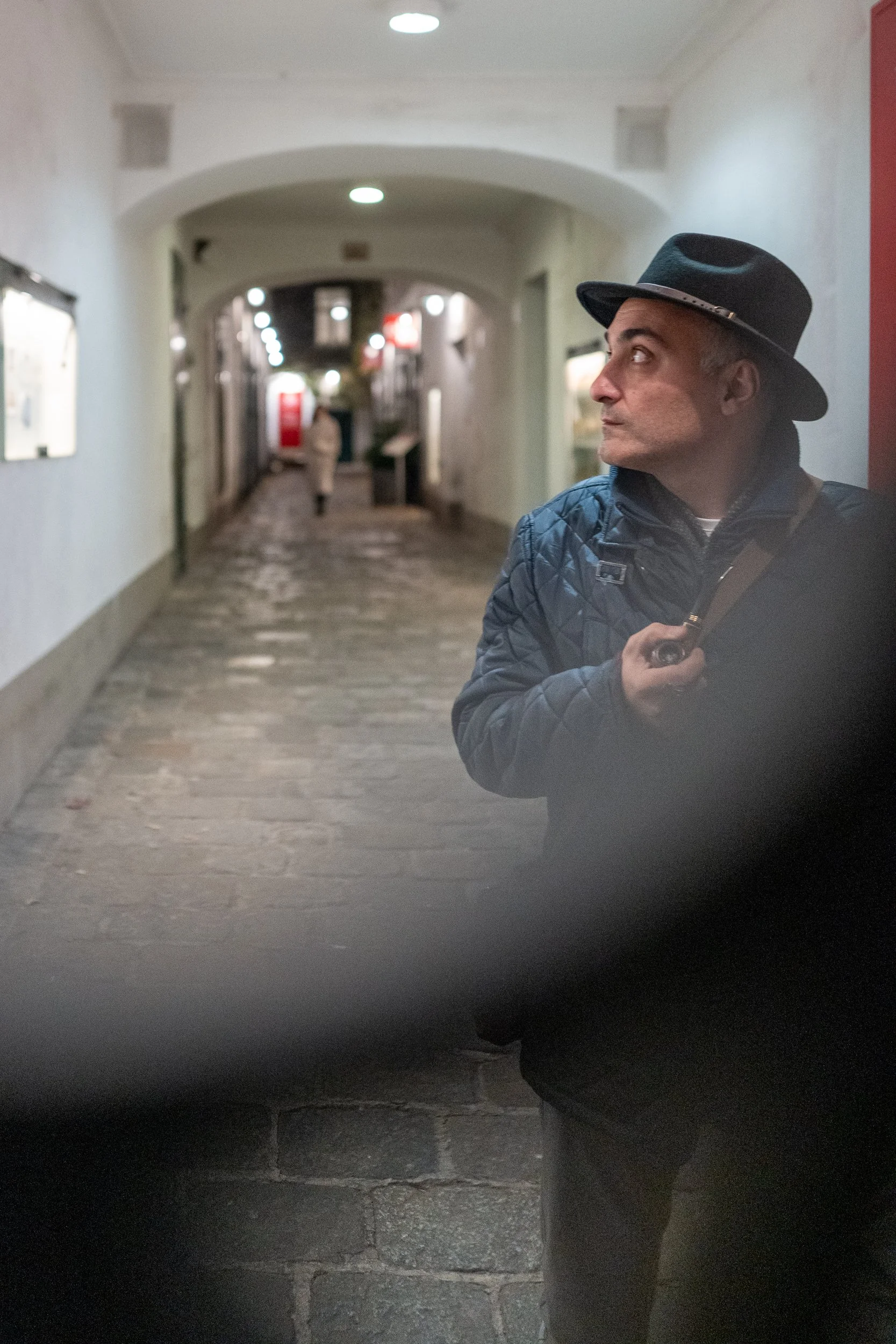 Un homme portant un chapeau noir et un manteau bleu est dans une galerie commerciale ou une zone piétonne, regardant à droite, tenant un appareil photo ou un téléphone dans la main.