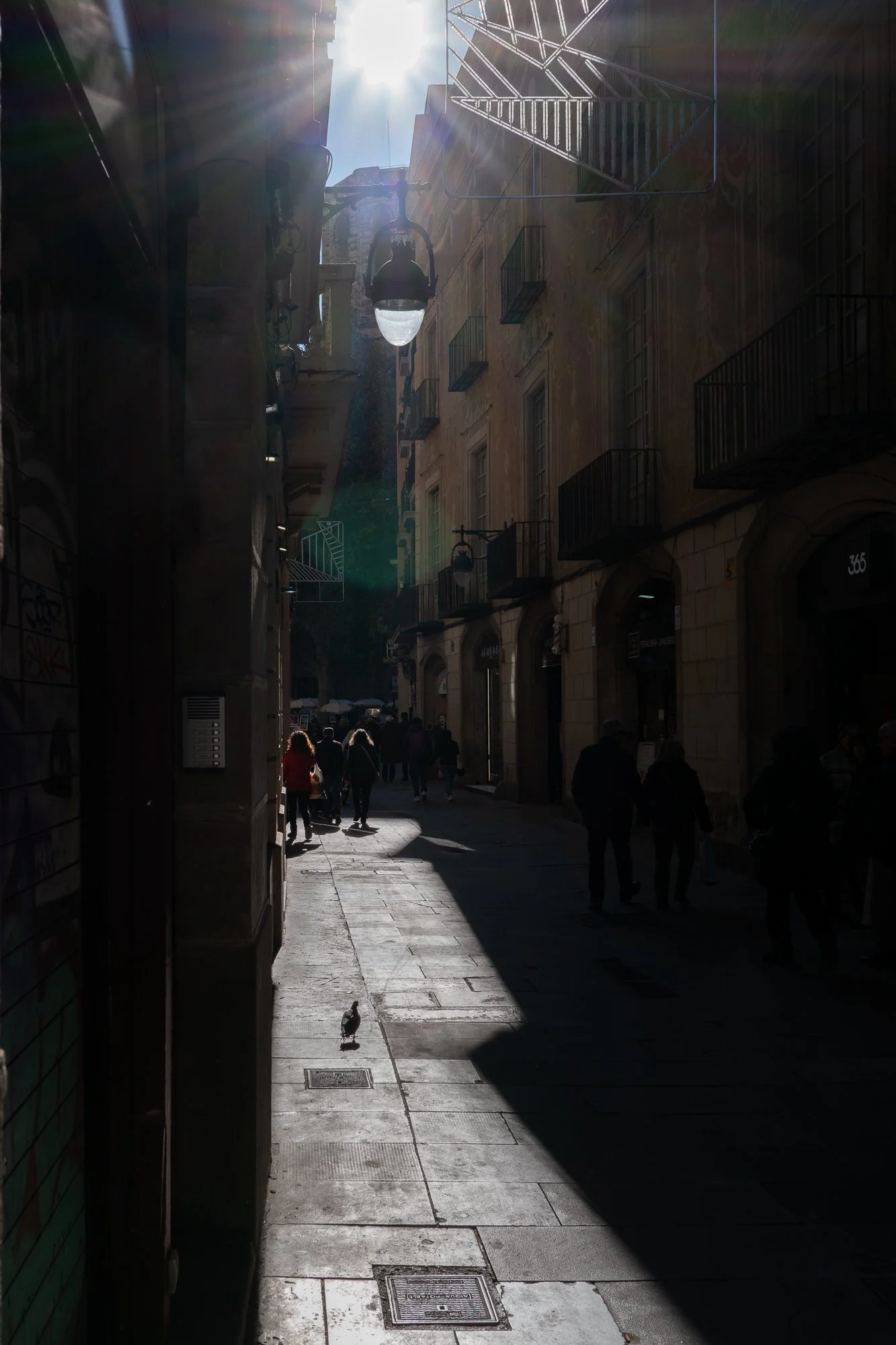 Rue étroite avec des bâtiments anciens, plusieurs personnes marchant, le soleil brillant fortement, ombres longues sur le sol, lampadaires suspendus