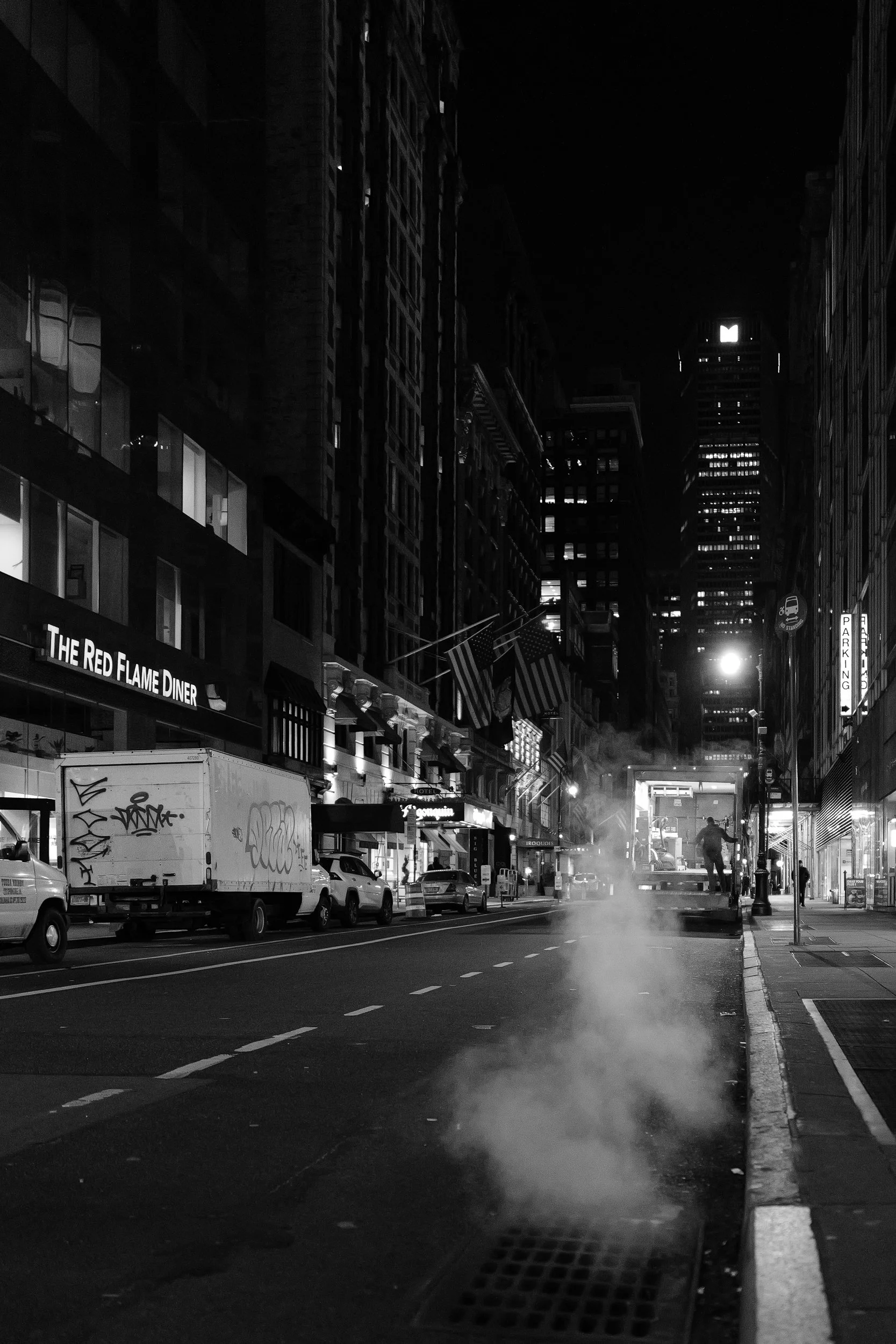Rue urbaine de nuit avec des voitures stationnées, un brumisateur ou vapeur s'échappant du trottoir, bâtiments avec fenêtres éclairées, enseignes lumineuses y compris 'The Red Flame Diner', drapeaux, et un gratte-ciel dans le ciel noir.