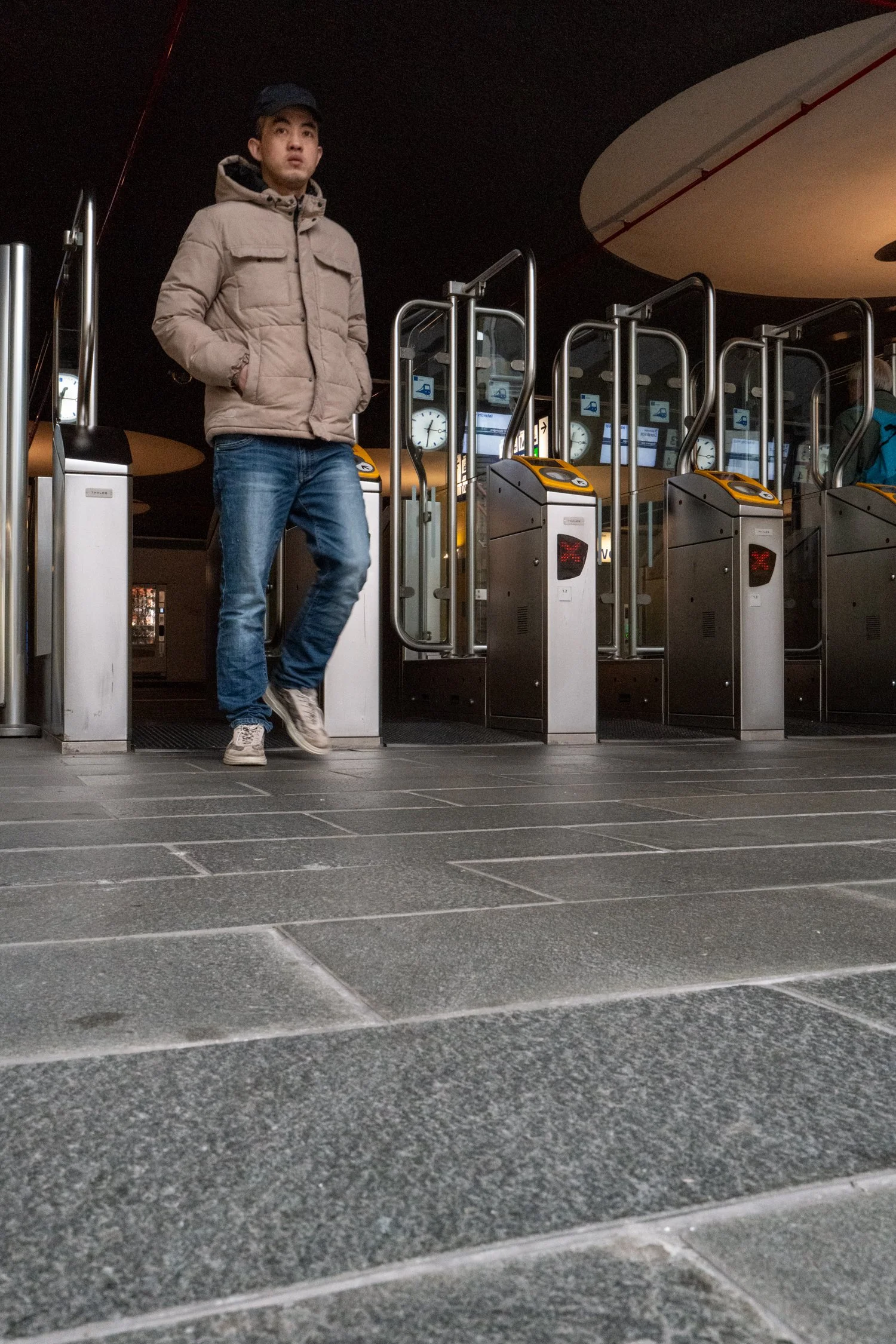 Un jeune homme debout à une entrée de station de métro, portant une veste beige et un pantalon en jean, avec une station de métro moderne en arrière-plan.