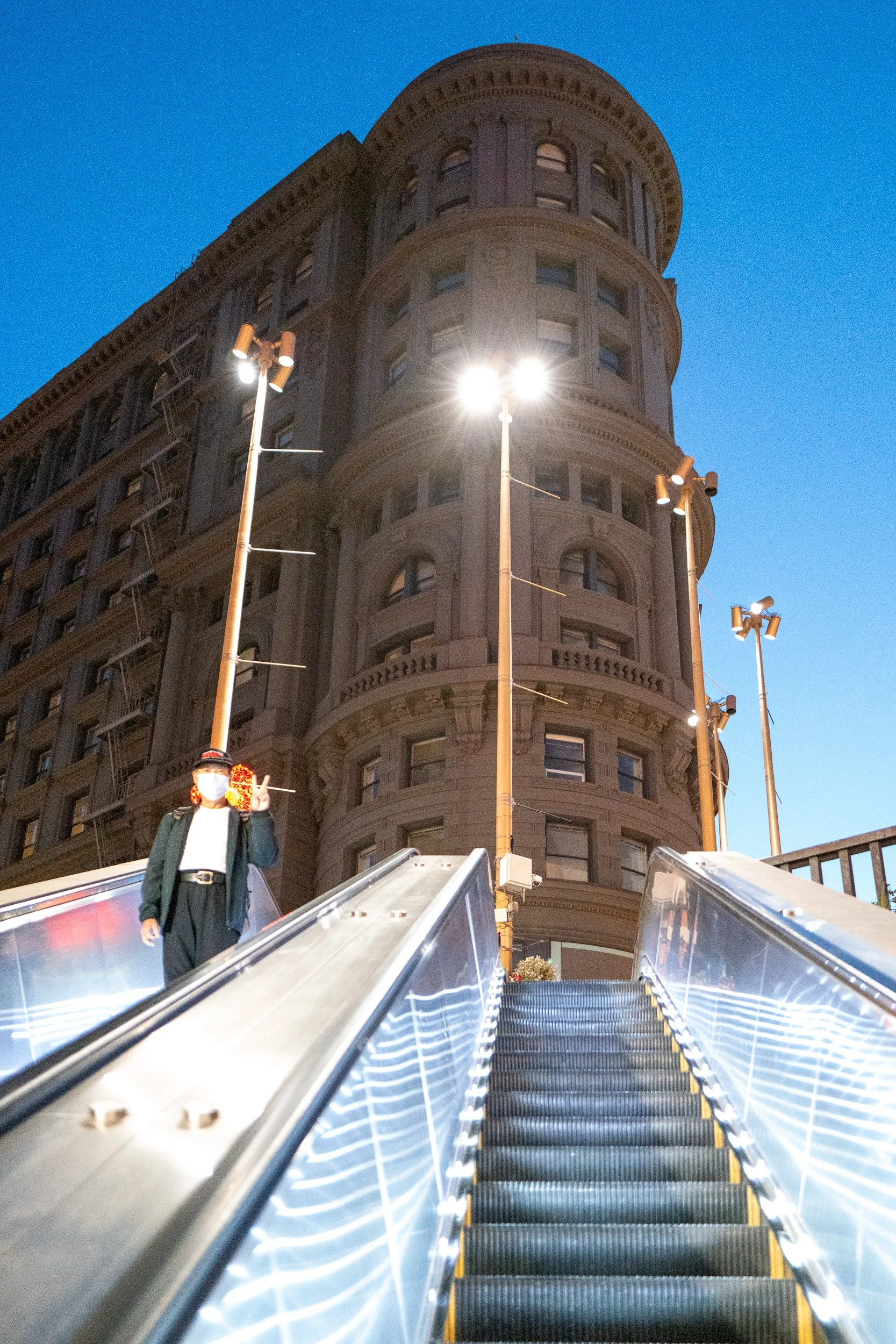 Une personne portant un masque, debout en haut d'un escalator en extérieur, avec un bâtiment ancien en arrière-plan, éclairée par des lampadaires nocturnes. Ciel bleu clair.