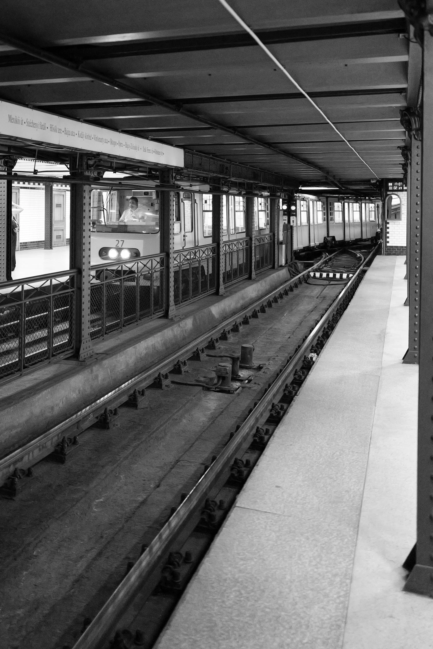 Un métro à quai avec un conducteur visible à travers la vitre, station souterraine japonais, image en noir et blanc.