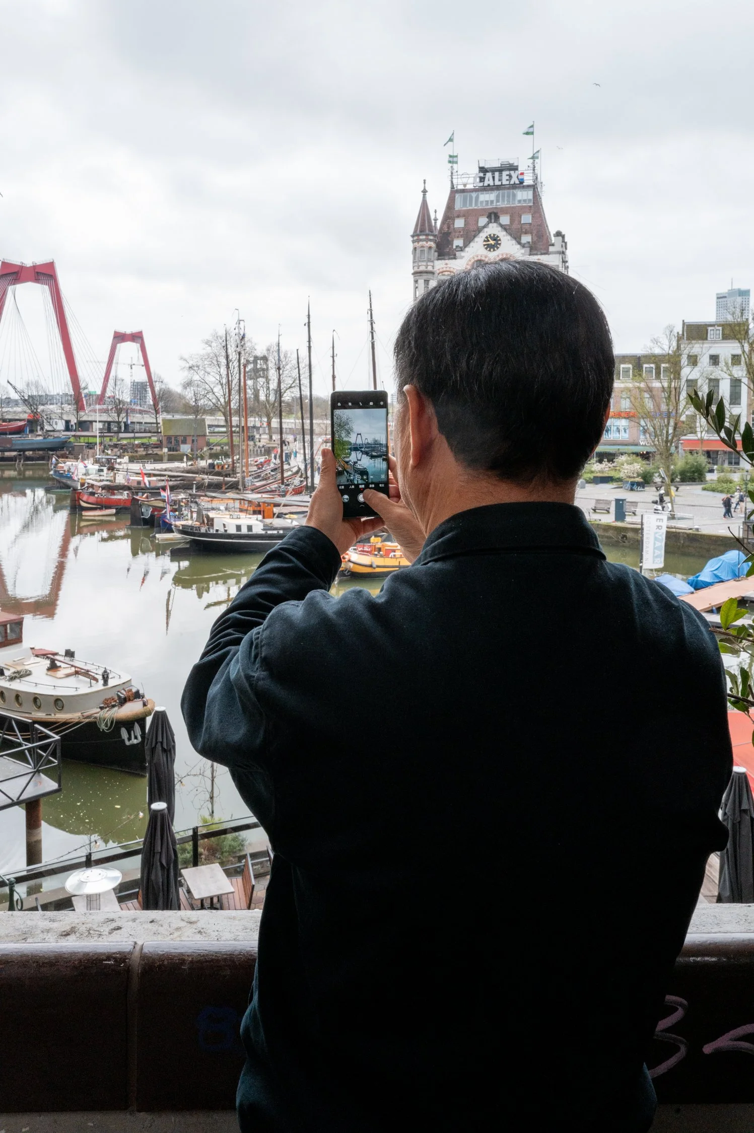 Une personne avec des cheveux courts, portant un vêtement noir, prend une photo avec son téléphone portable d'un paysage maritime avec des bateaux, des bâtiments historiques, une horloge et des parcs au bord de l'eau, sous un ciel gris.