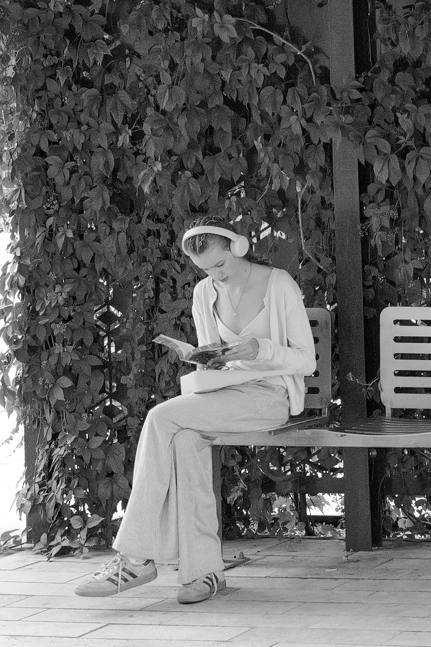 Une jeune femme assise sur une chaise, portant des écouteurs, lisant un livre, avec un mur recouvert de feuillage en arrière-plan.