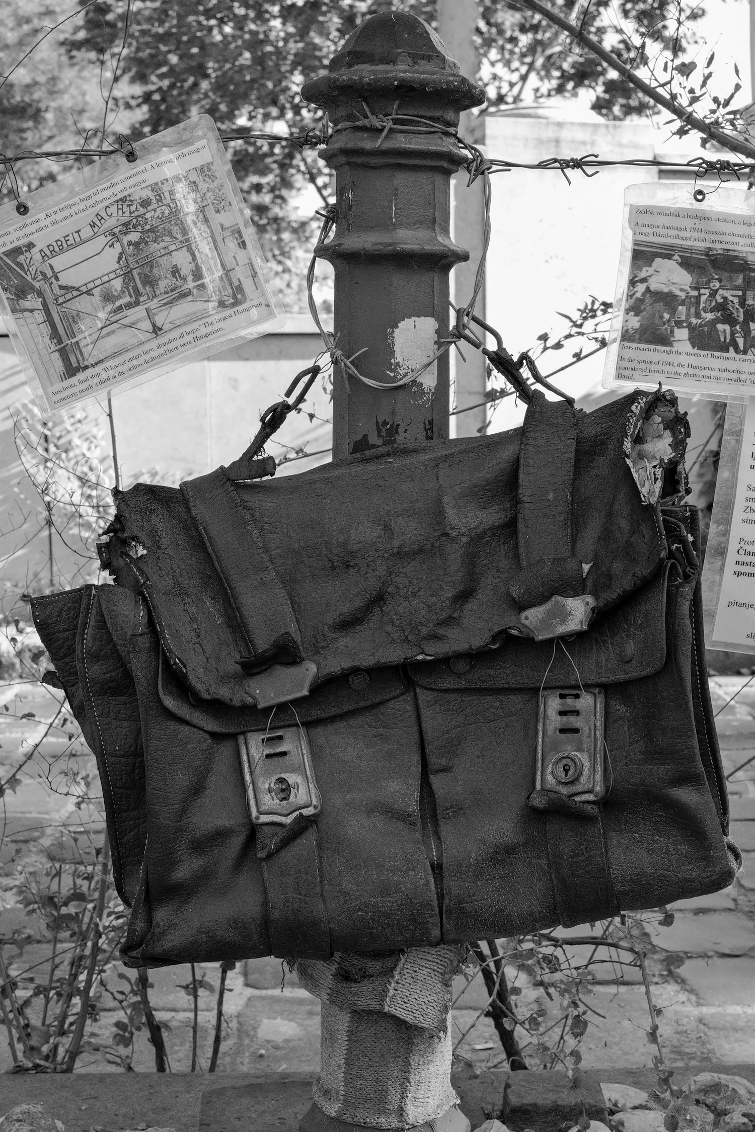 Une boîte aux lettres ancienne en métal fixée à un poteau en béton, avec des documents accrochés à des fils de barbelés à l'arrière-plan, dans un environnement extérieur.