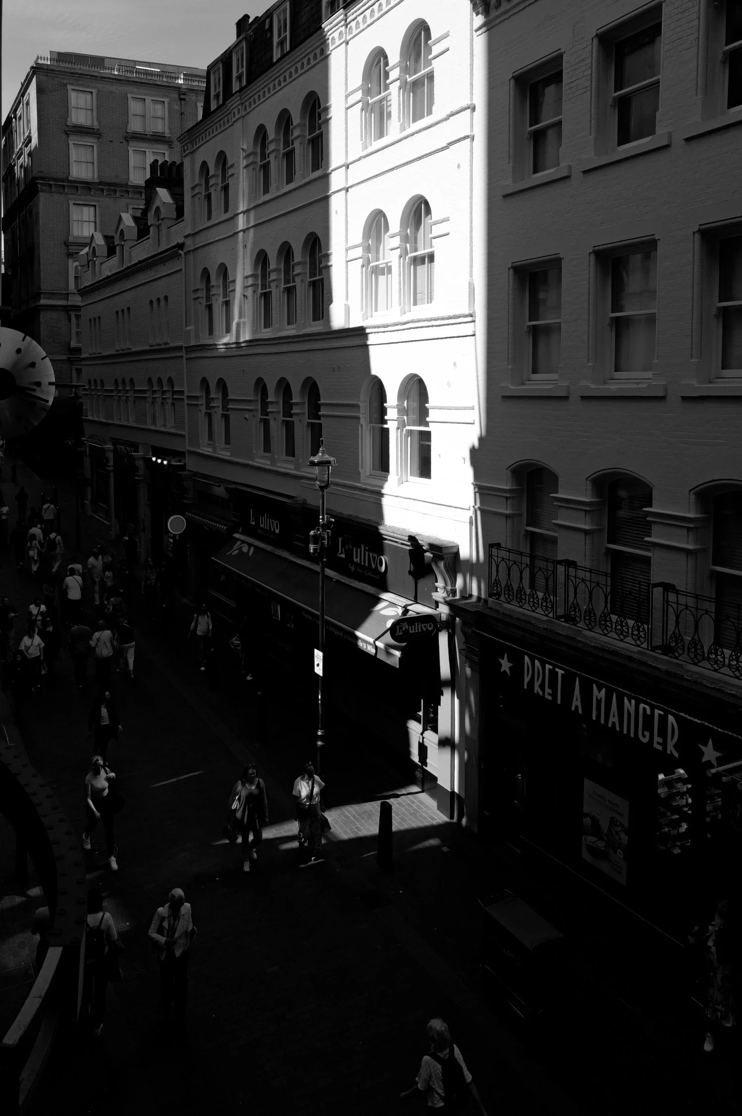 Une rue urbaine avec des bâtiments à plusieurs étages, des passants marchant sur le trottoir, une boutique avec un grand affichage et un lampadaire en fer forgé. L'image est en noir et blanc.