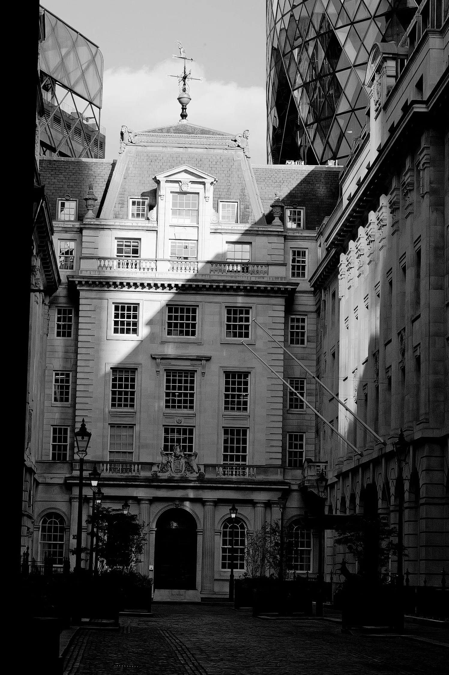Vue d'un bâtiment historique moderne et ancien sous un ciel nuageux, avec une rue pavée en premier plan et des lampadaires, en noir et blanc.