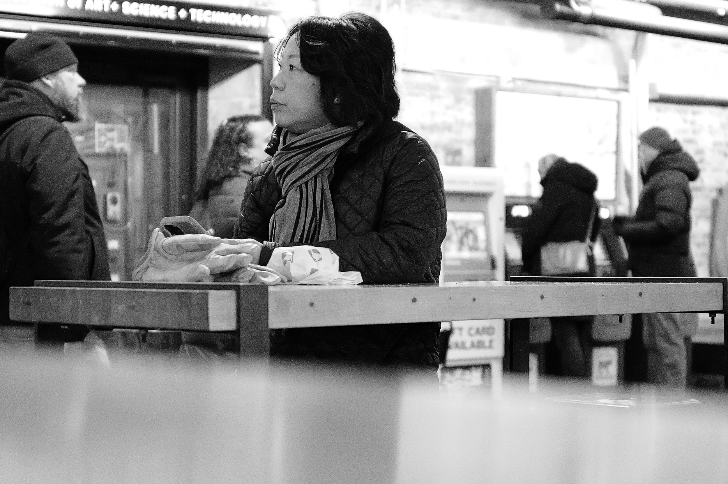Une femme debout à une table, regardant une téléphone portable, dans un espace intérieur avec plusieurs personnes en arrière-plan.
