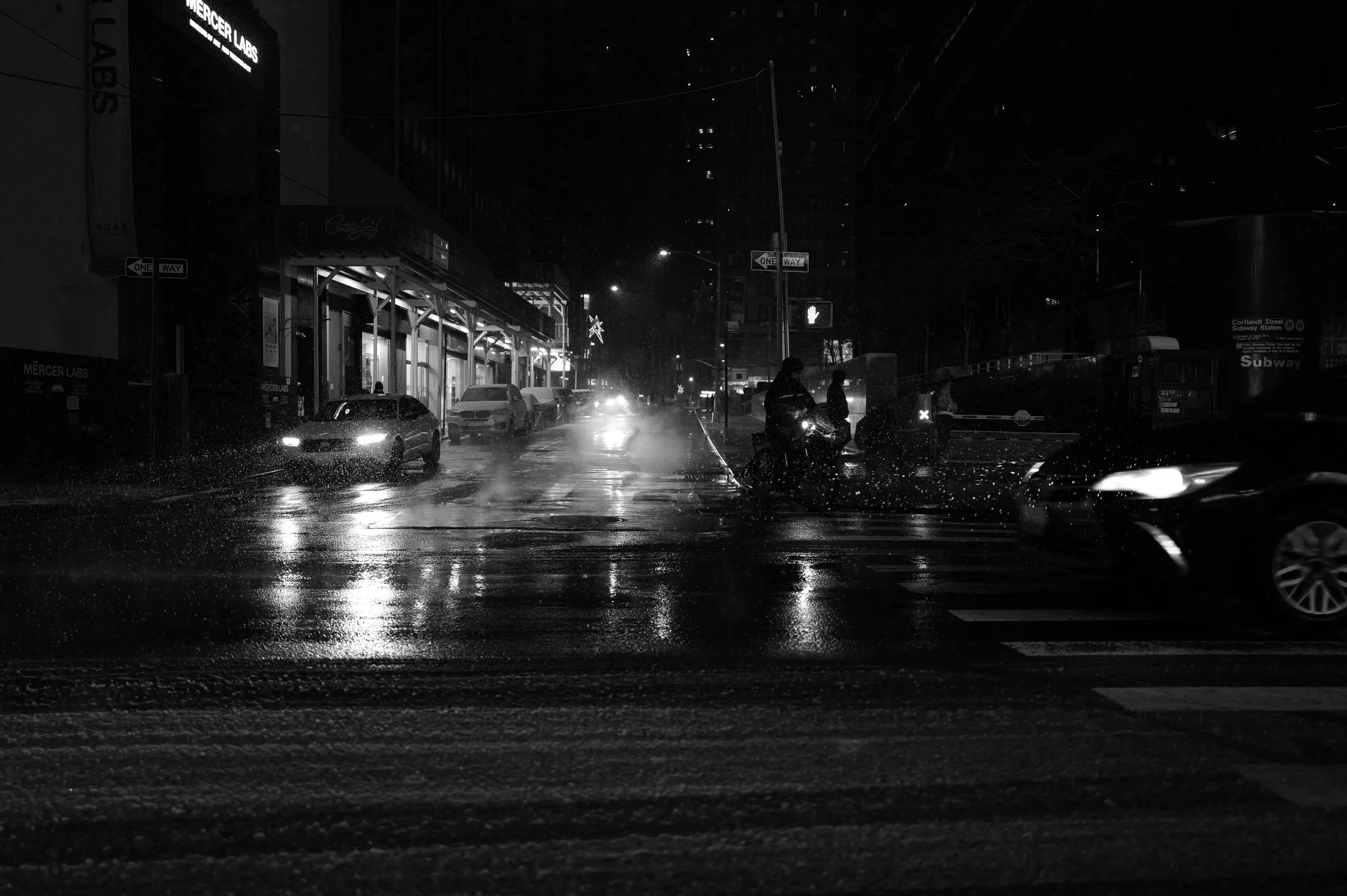 Une rue urbaine la nuit sous la pluie, avec des voitures, des scooters, des passants avec parapluie, et des enseignes lumineuses, en noir et blanc.