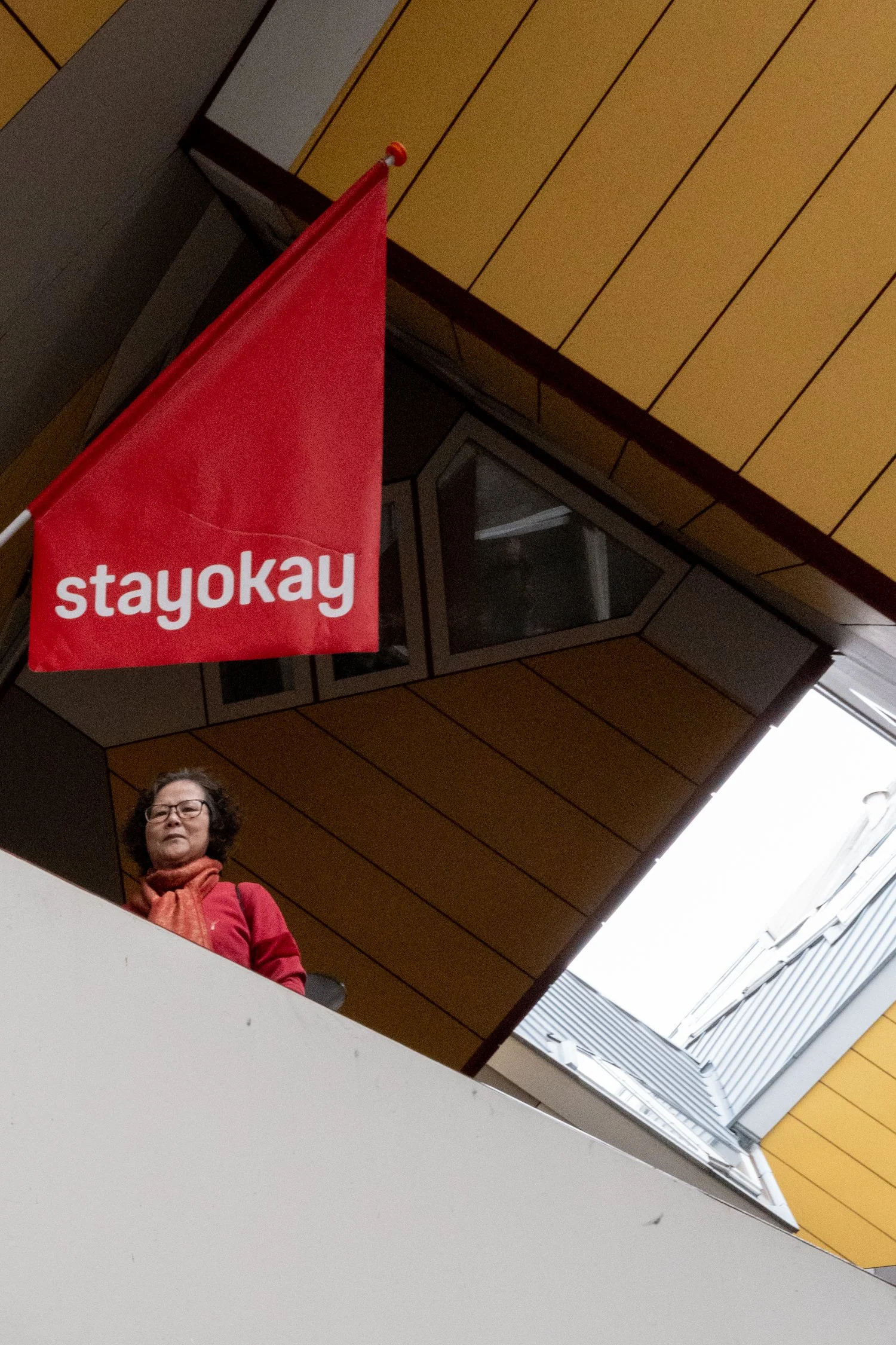 Une femme portant des lunettes, une écharpe orange et un pull rouge, regardant vers l'avant, se tient dans un bâtiment moderne avec un plafond en bois, une lucarne et un drapeau rouge avec le mot 'stayokay'