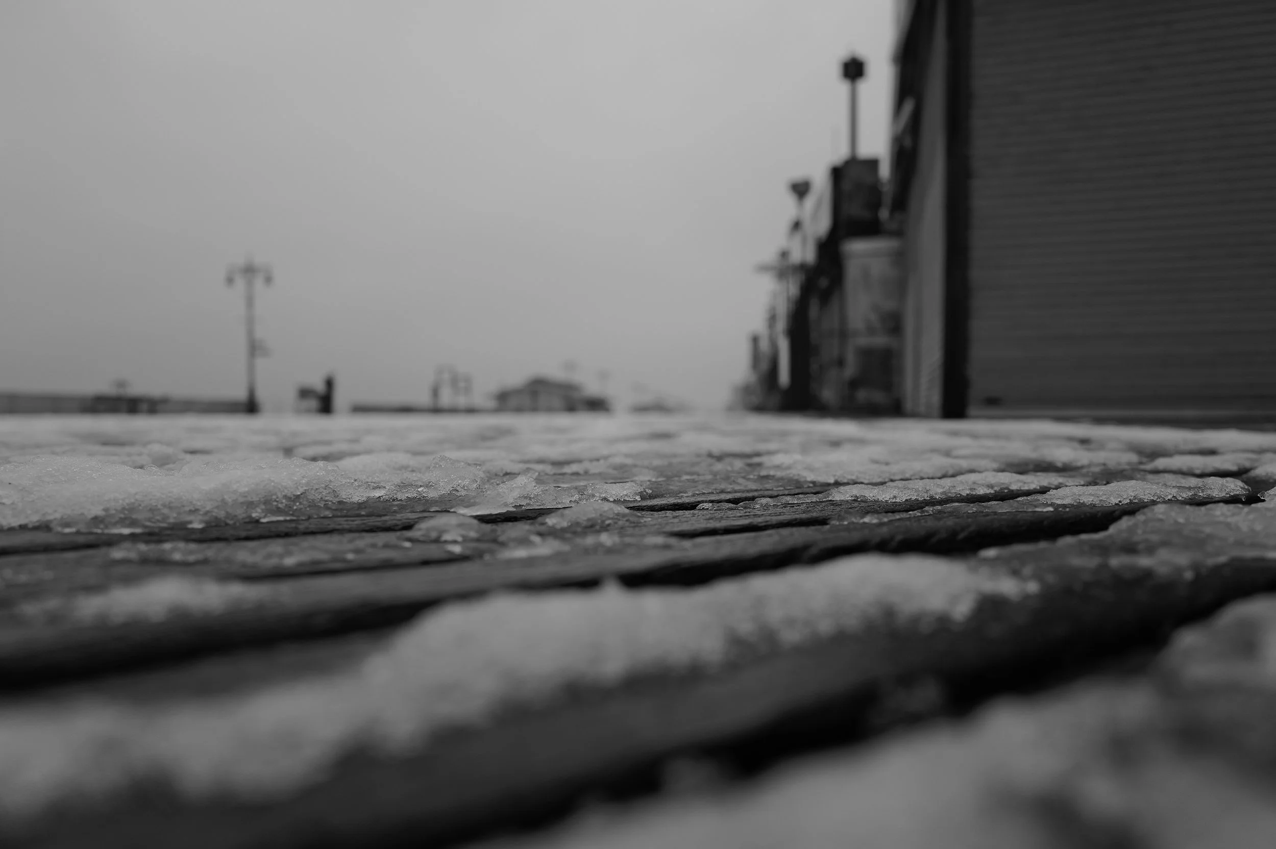 Vue au ras du sol d'une surface couverte de glace ou de neige, avec des bâtiments et des pylônes électriques flous en arrière-plan, sous un ciel nuageux.