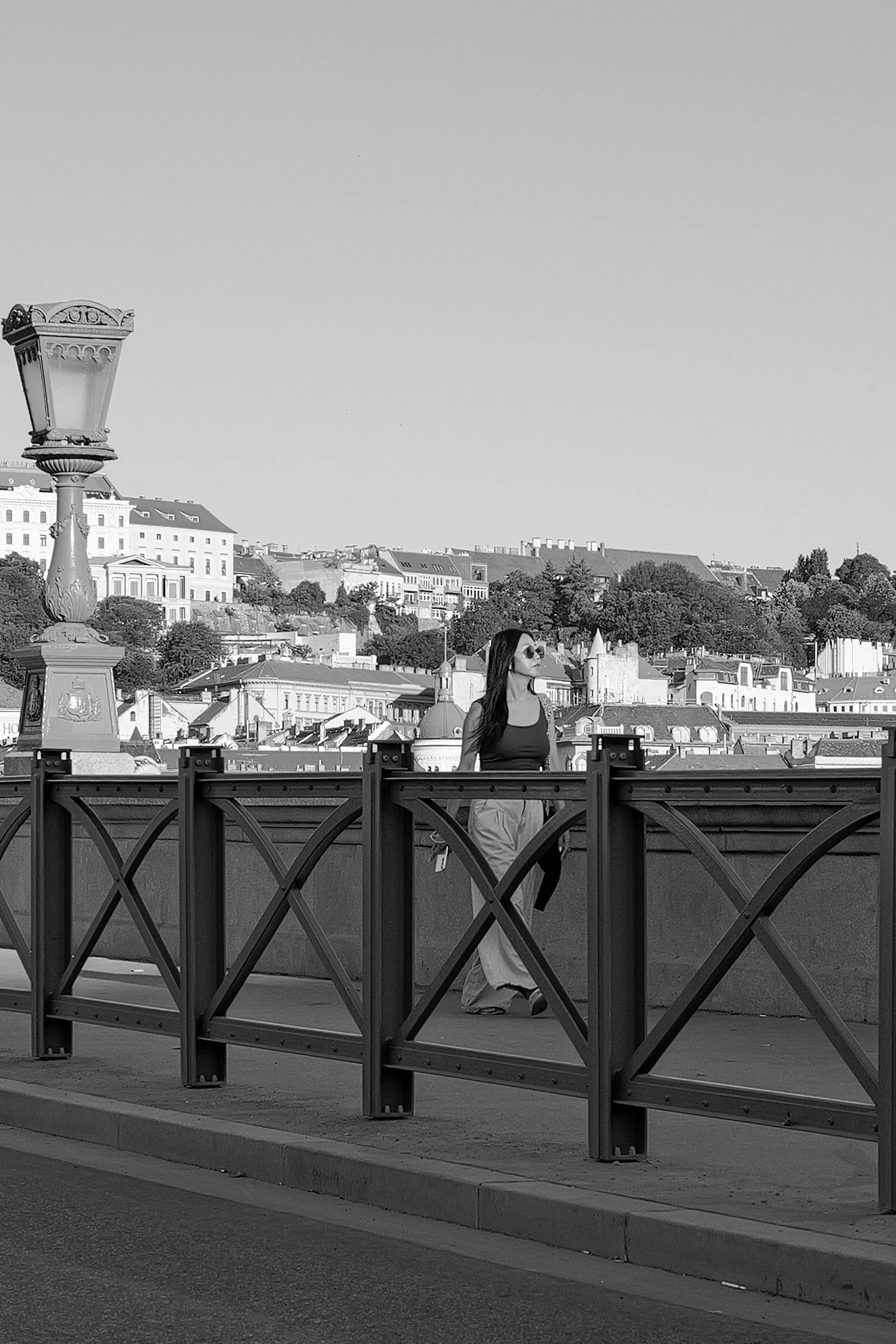Une femme portant des lunettes de soleil et un chapeau, marchant sur un pont avec une rambarde en métal, avec une ville en arrière-plan. La photo est en noir et blanc.