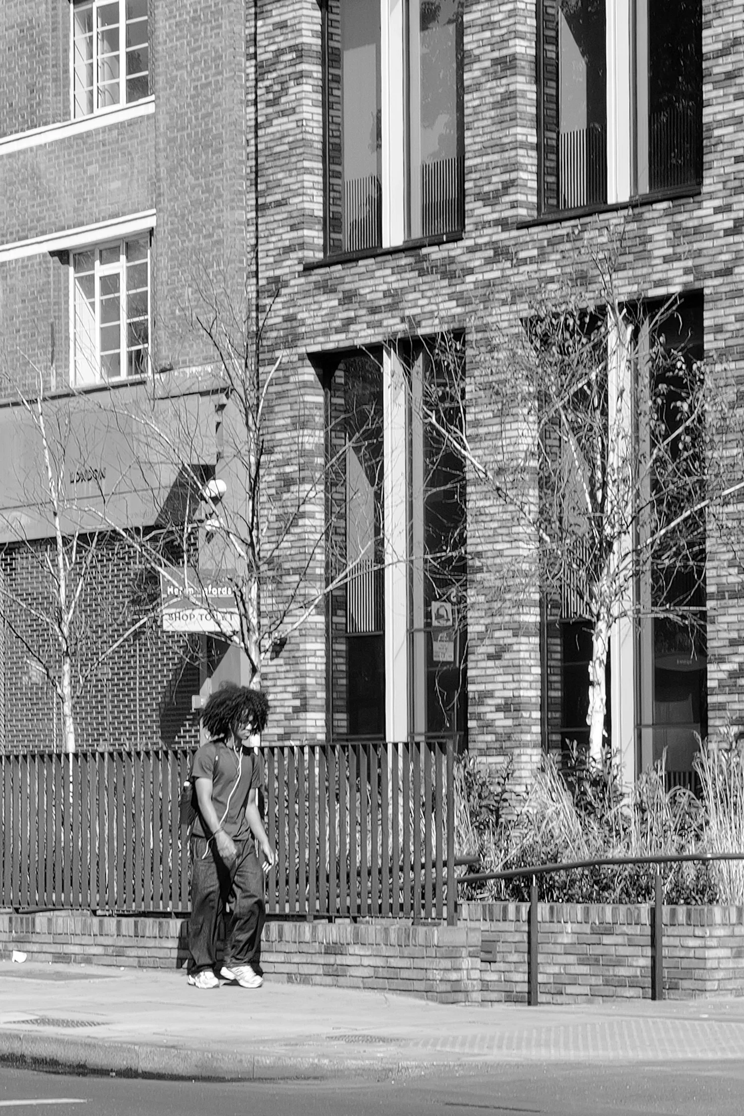 Une personne avec des cheveux frisés portant un sweatshirt à capuche et un pantalon décontracté marche sur le trottoir, en arrière-plan, un bâtiment en briques avec plusieurs fenêtres et un garde-corps en métal.