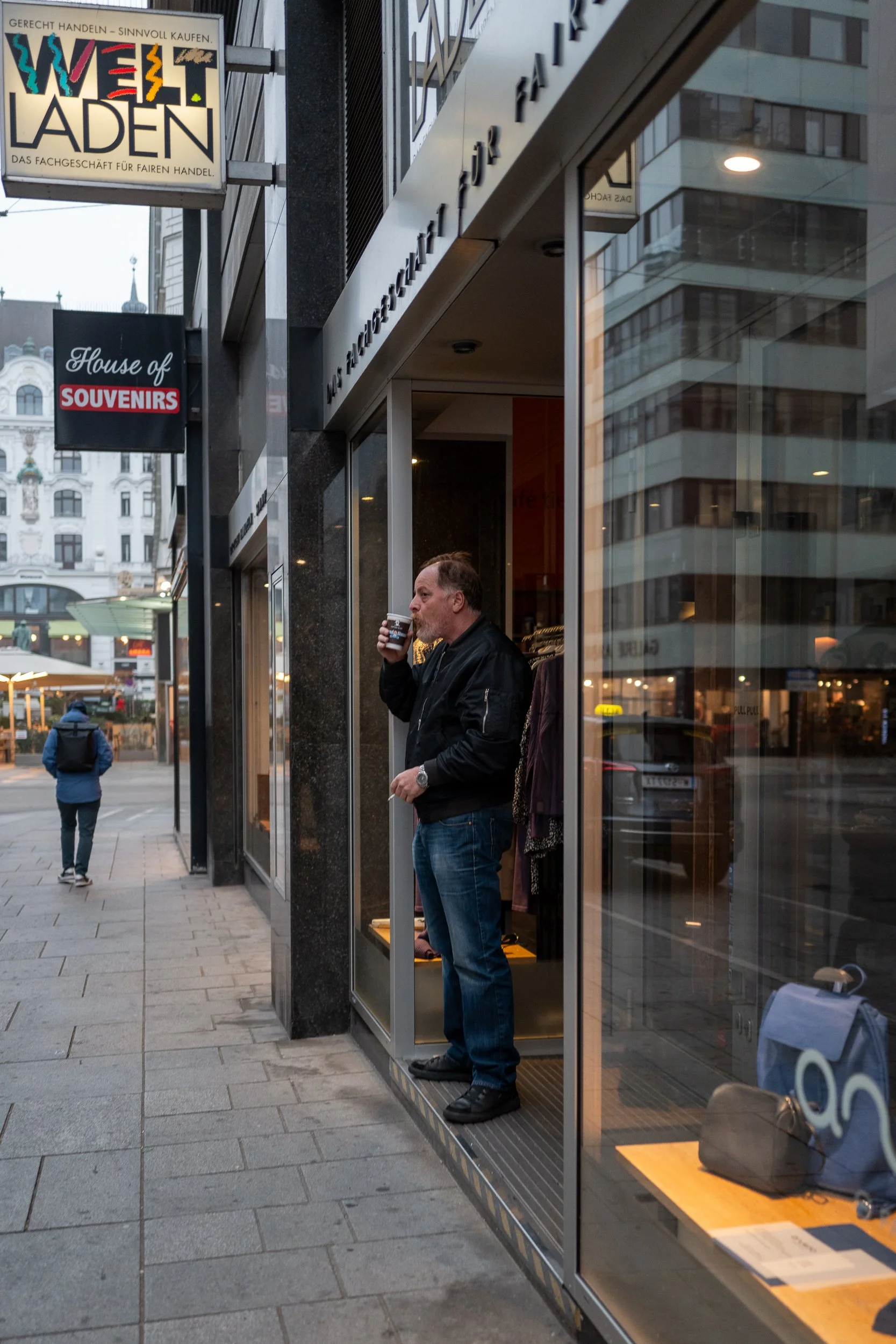 Un homme debout à la sortie d'une boutique, buvant une boisson, avec une vitrine en verre et des immeubles en arrière-plan, dans une ville urbaine.