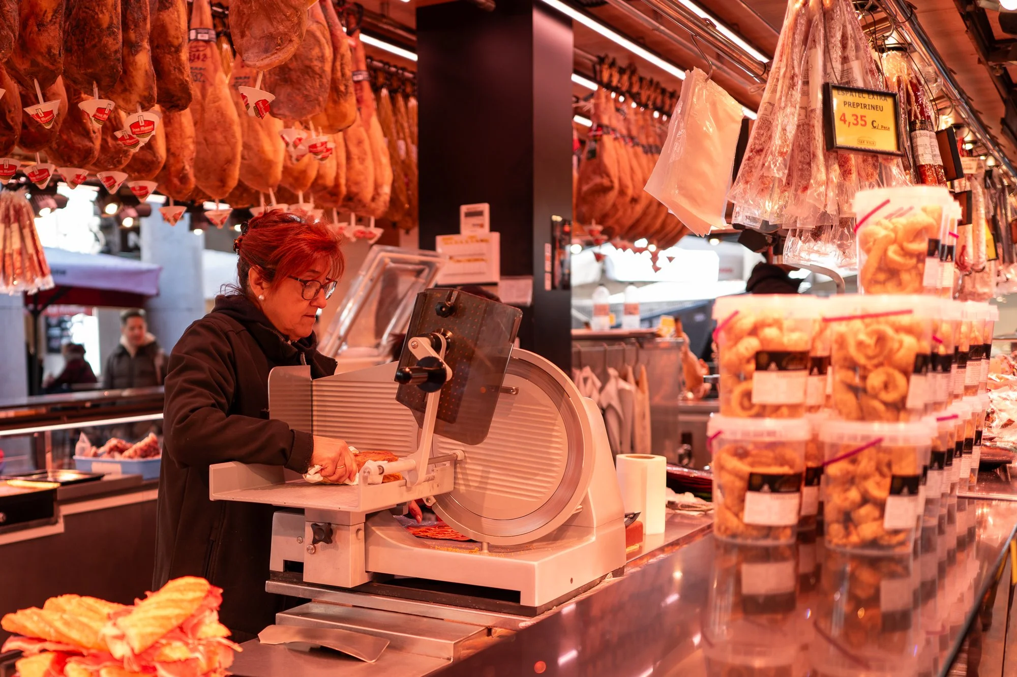 Une femme coupe de la charcuterie dans une boucherie ou un marché, avec des jambons suspendus et des paquets de produits emballés sur le comptoir.