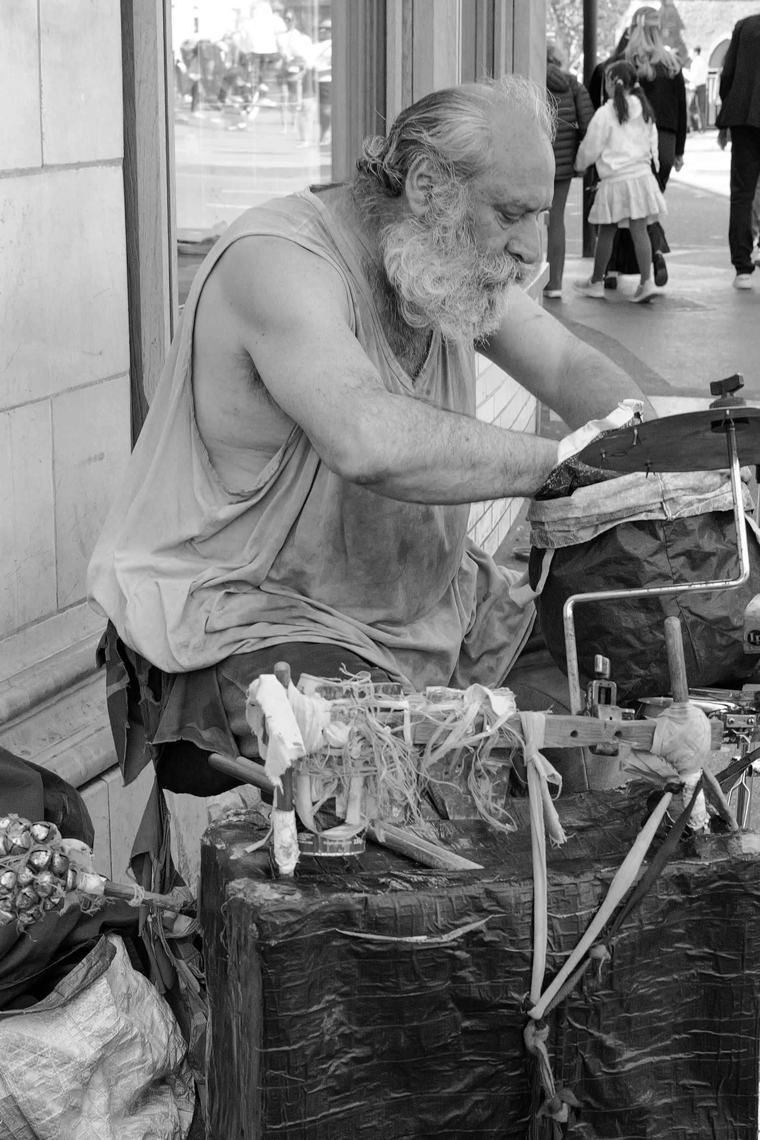 Un homme avec une barbe grise joue de la musique dans la rue, assis devant un stand avec des objets divers, entouré de passants, en noir et blanc.