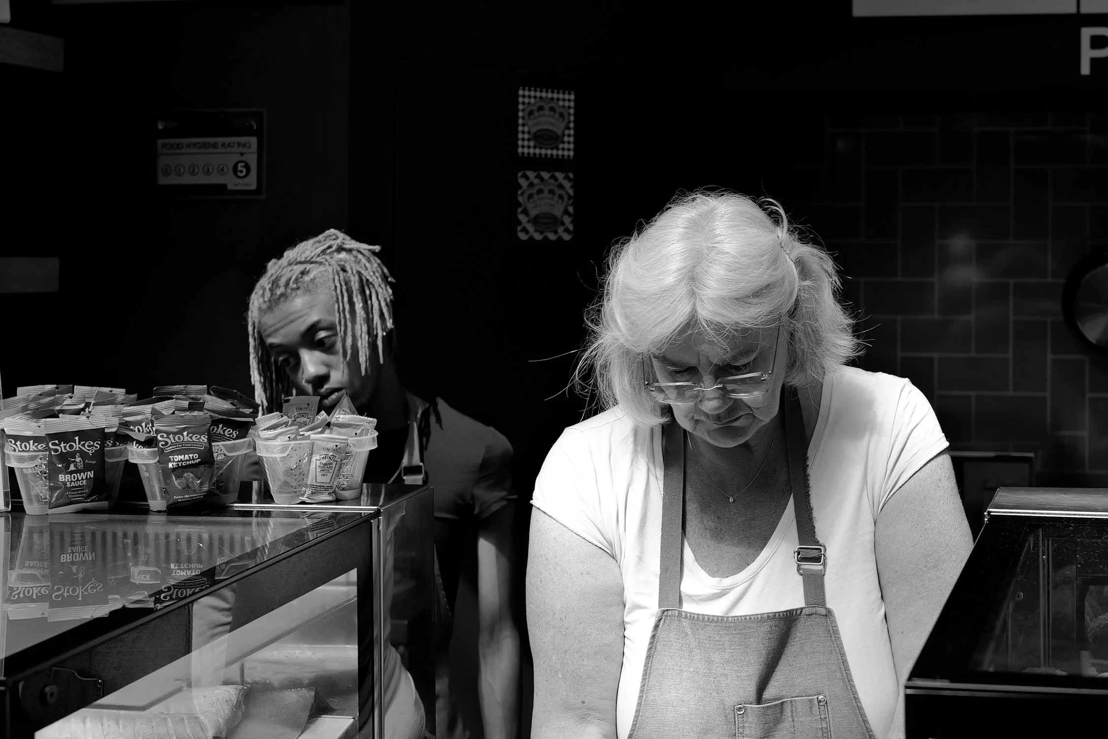 Deux femmes, une avec des cheveux frisés blancs portée en gilet blanc et une autre avec des cheveux longs frisottés, travaillent derrière un comptoir dans un environnement de restauration rapide. Elles se concentrent sur leur tâche, probablement en t