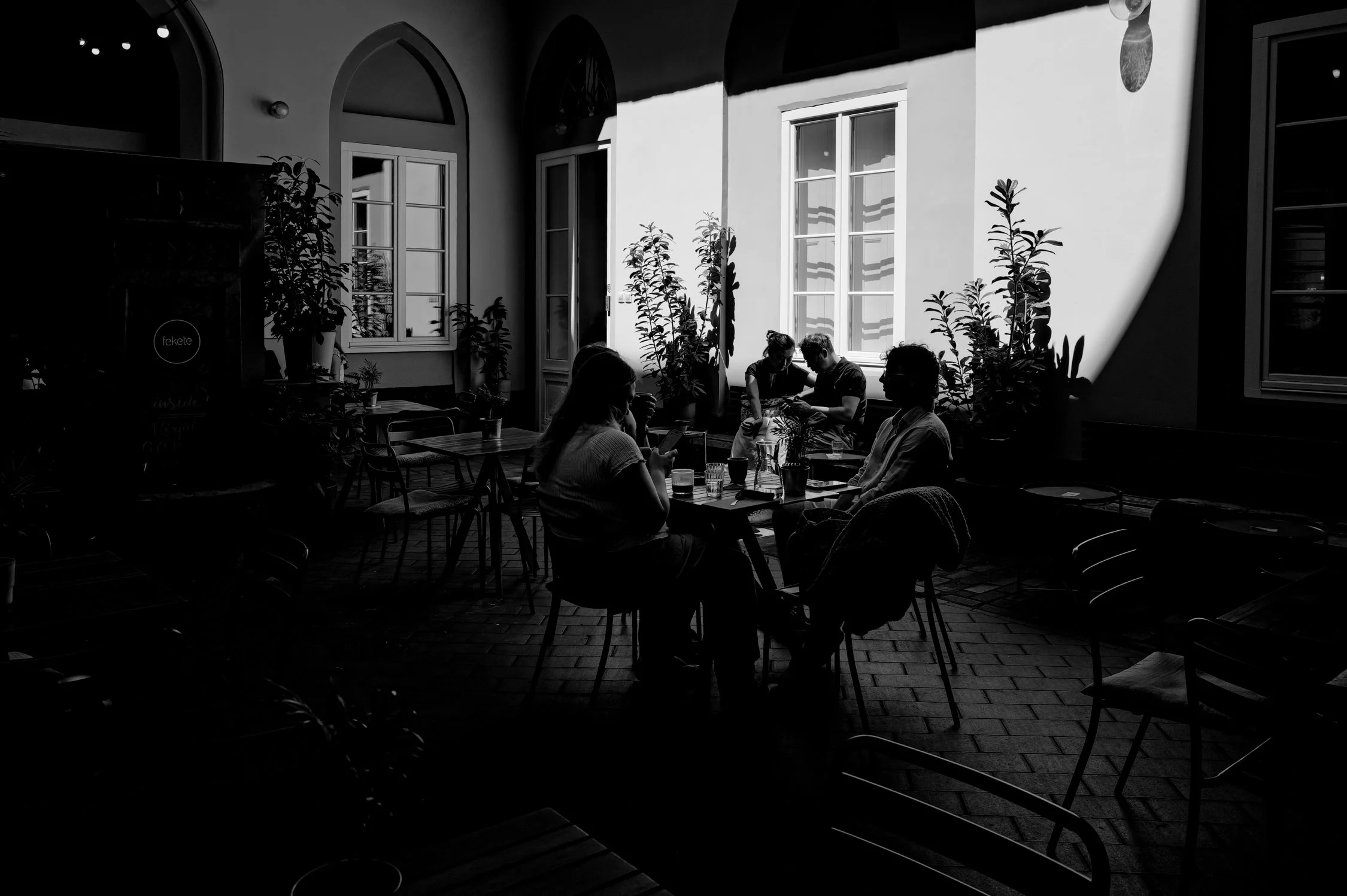 Salle à manger ou café avec plusieurs personnes assises à une table, éclairée par la lumière naturelle venant de grandes fenêtres avec des plantes