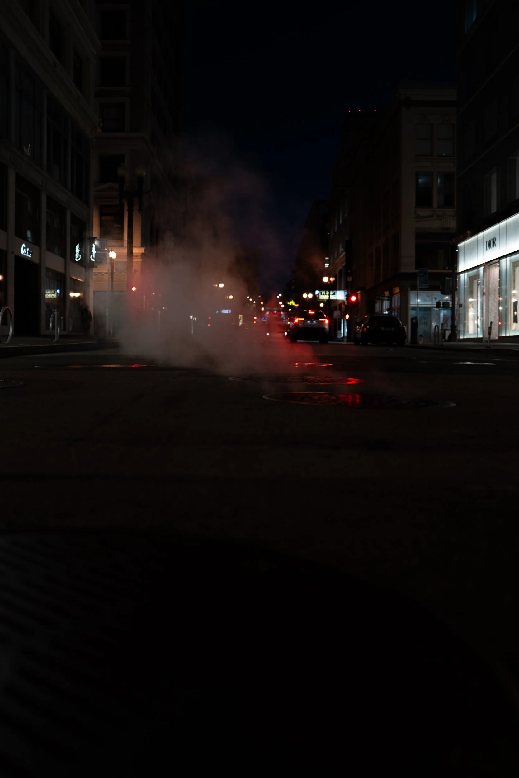 Une rue urbaine la nuit avec de la vapeur ou de la fumée qui s'élève du sol, entourée de bâtiments avec des vitrines éclairées et quelques voitures Stationnées.