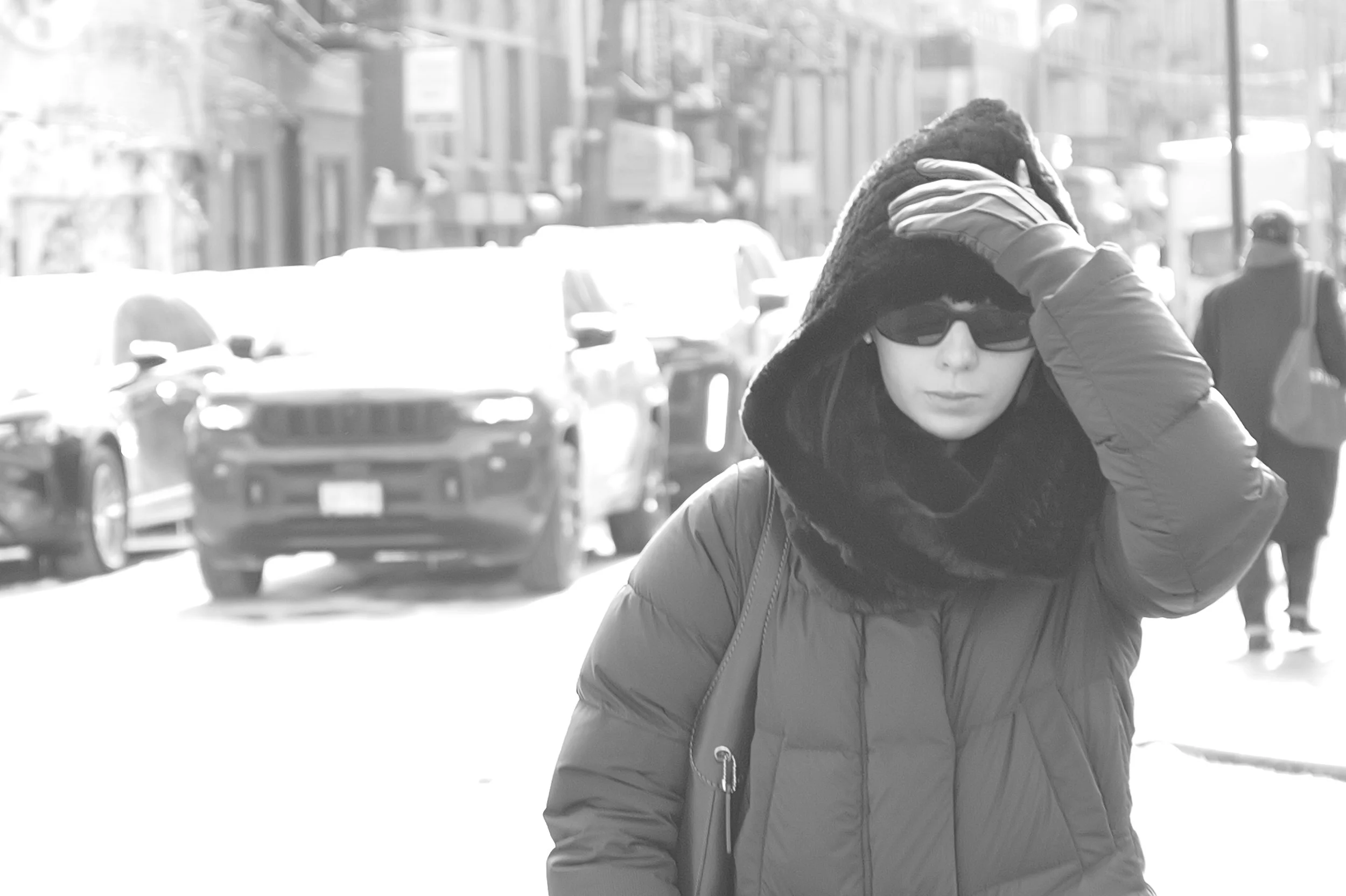 Femme portant un manteau à capuche avec cache-oreilles, portant des lunettes de soleil, dans une rue urbaine en hiver.