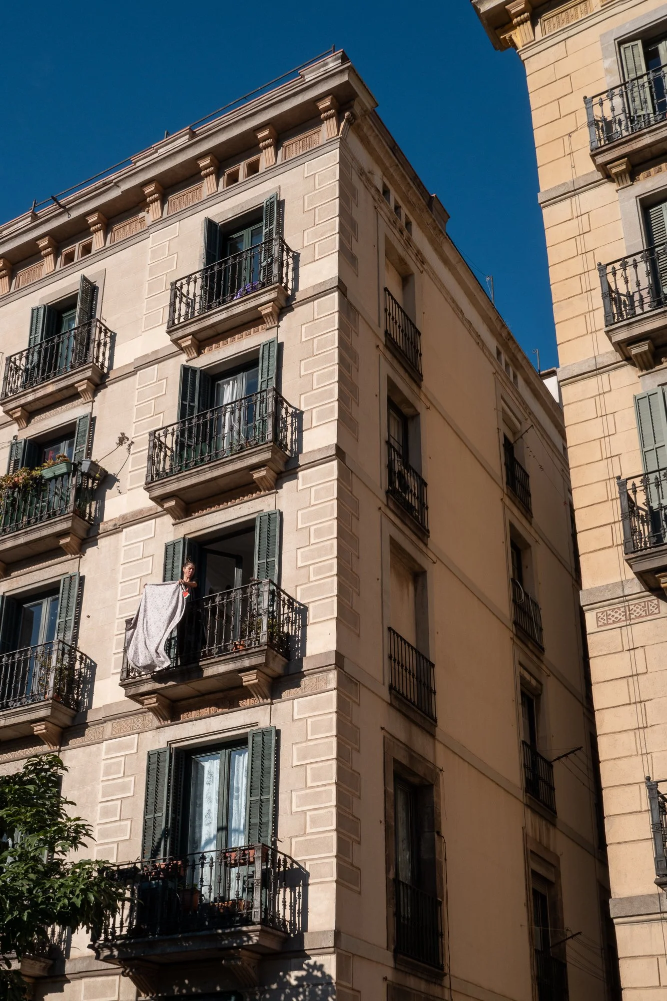 Un immeuble résidentiel en pierre beige avec plusieurs balcons en fer forgé noir et des fenêtres avec volets verts. Une personne est sur un balcon, suspendant une pièce de linge à sécher. Le ciel est bleu clair, ensoleillé.