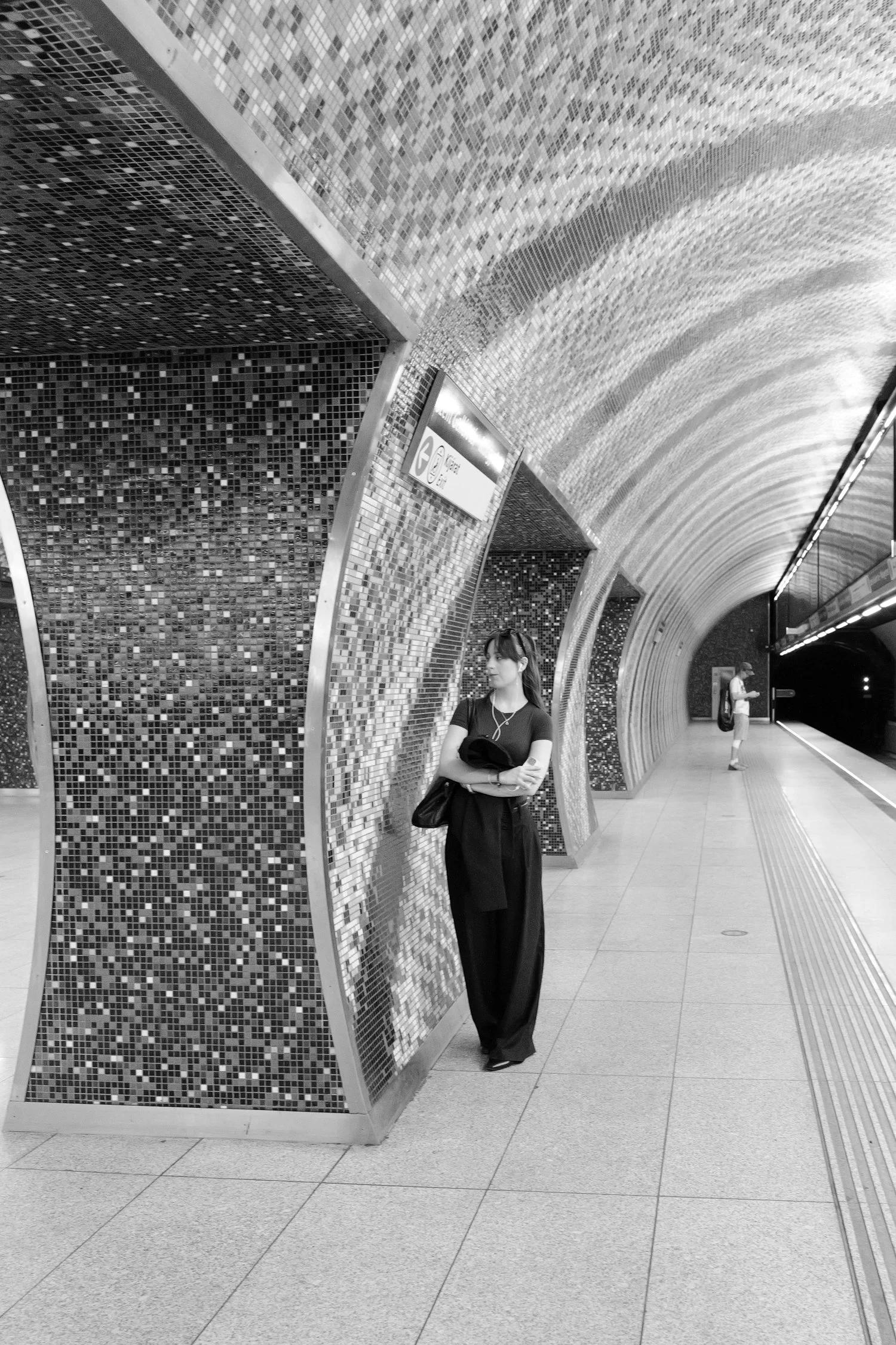 Deux femmes debout dans une station de métro, à côté d'une structure en mosaïque, l'une portant une robe longue noire et l'autre un t-shirt blanc et un sac à dos, l'atmosphère est calme.