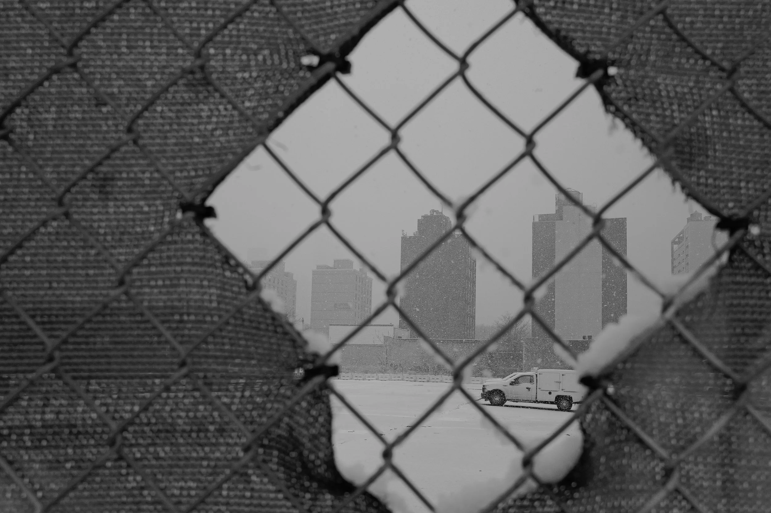 Une vue d'une ville enneigée prise à travers un trou dans une clôture en wire mesh, avec des bâtiments de hautes tours et un véhicule blanc sur la neige.