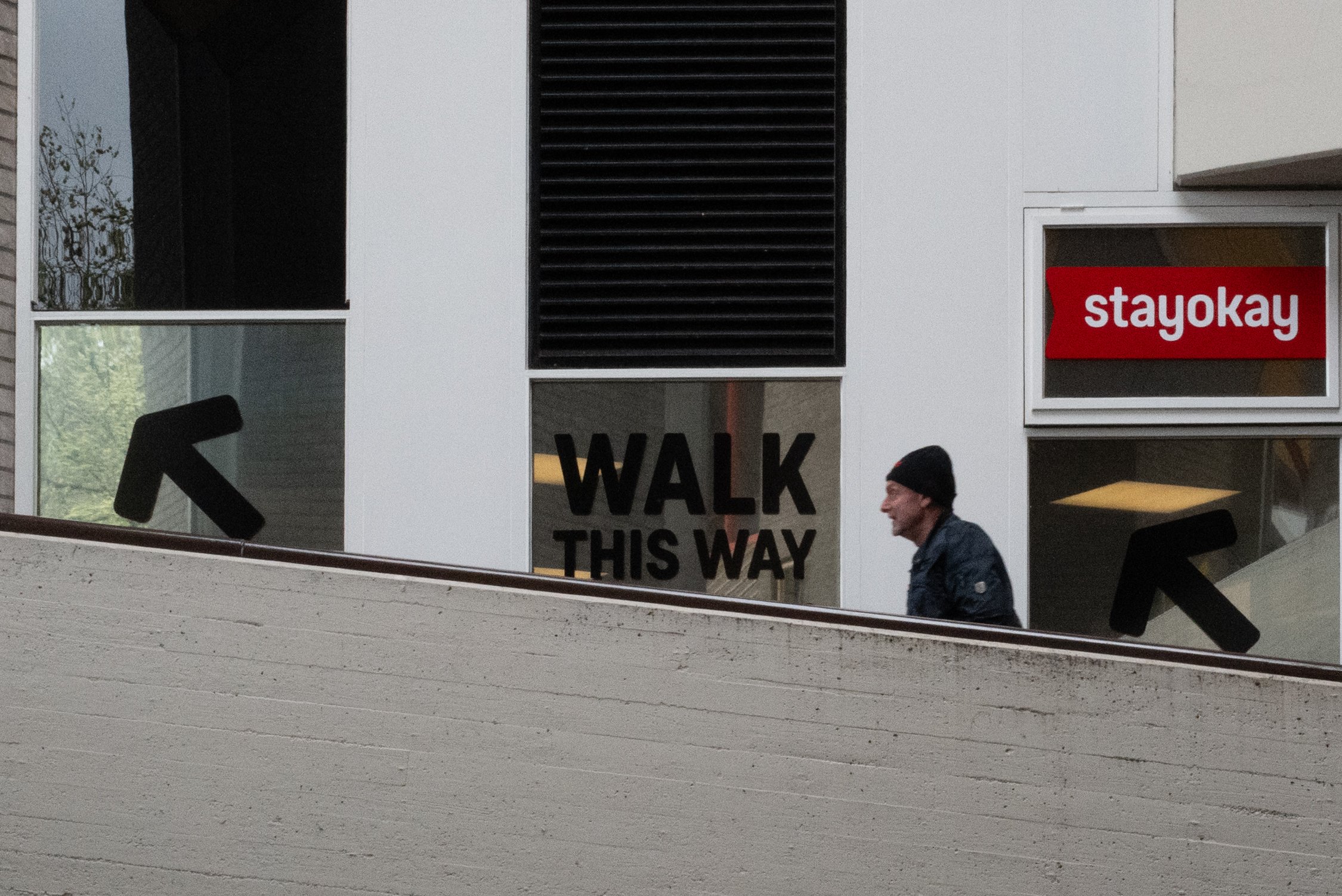 Une façade d'un bâtiment moderne avec plusieurs fenêtres, une ventilation noire, un panneau rouge avec l'inscription "stayokay", un panneau indiquant "WALK THIS WAY" en noir, et deux personnes marchant à proximité.