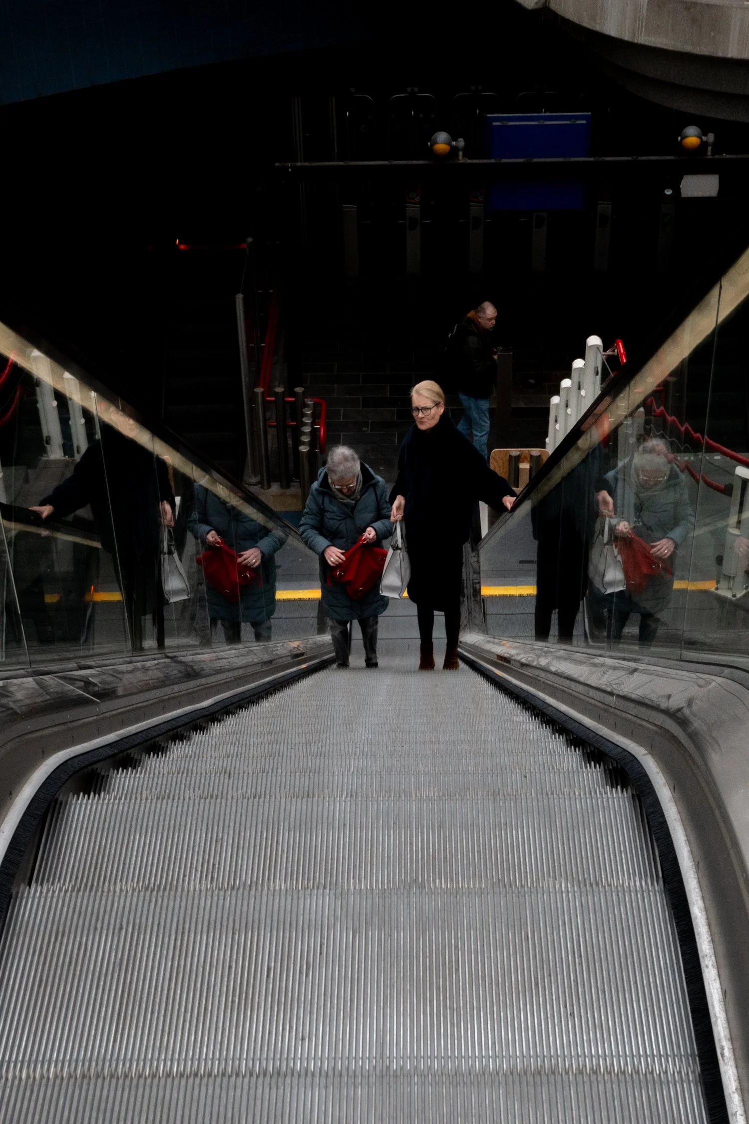 Groupe de personnes montant à pied une escalator dans une station souterraine, avec un reflet dans la vitre.