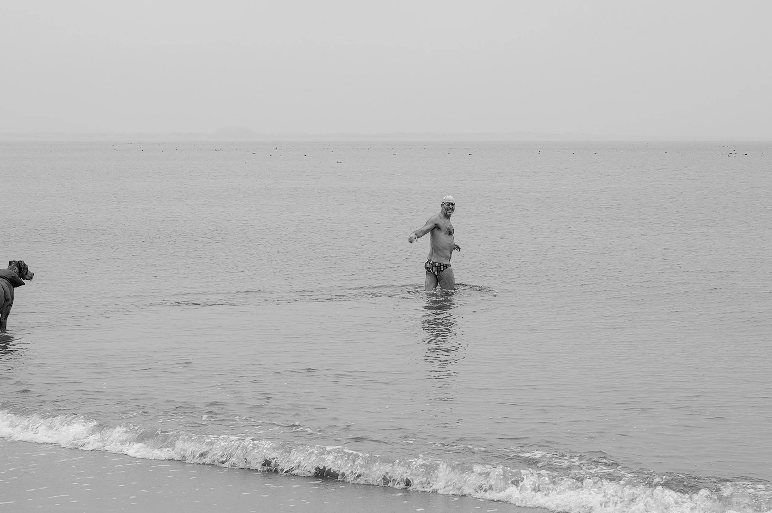 Un homme portant un maillot de bain à motifs, souriant, se tenait dans la mer, avec un chien sur la plage à gauche. L'image est en noir et blanc.
