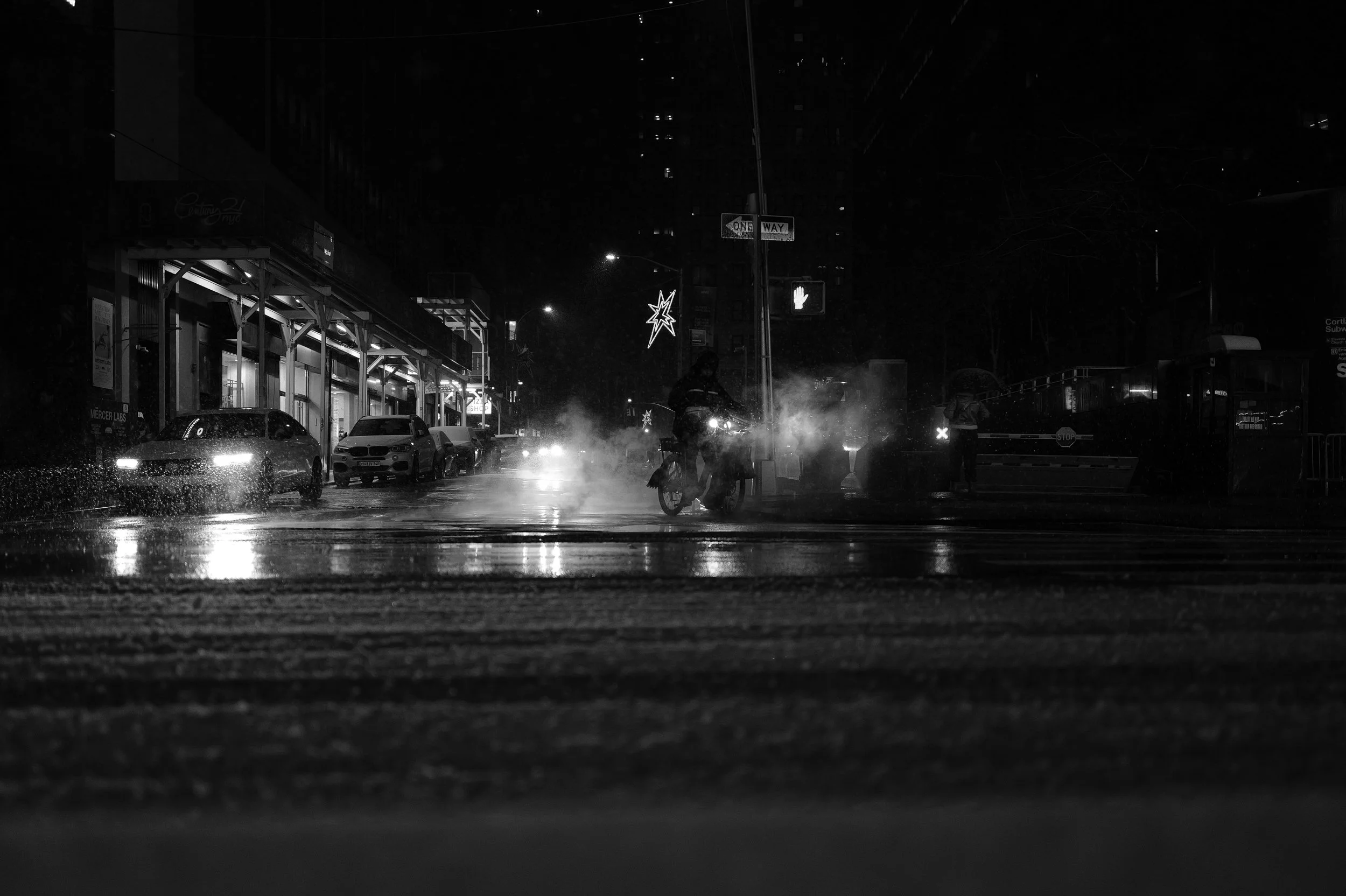 Une rue urbaine la nuit, avec des voitures stationnées, un cycliste, et une personne sous un parapluie. La scène est sombre, éclairée par des lumières de rue et des décorations lumineuses.
