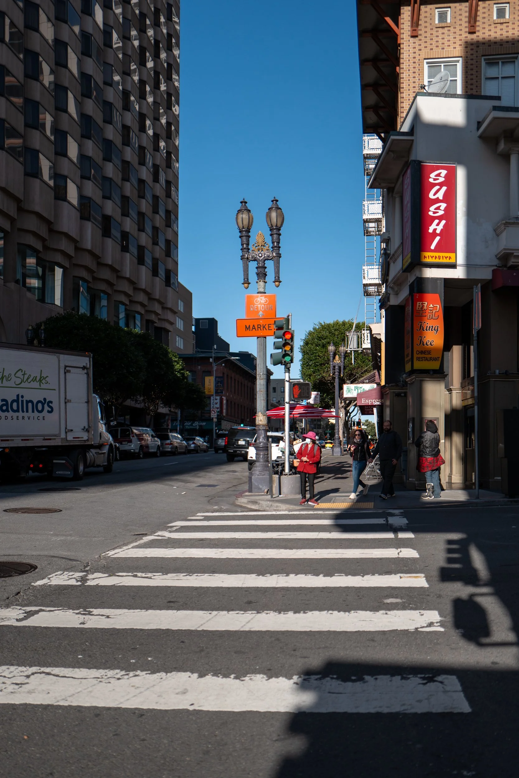 Rue urbaine avec un passage pour piétons, plusieurs piétons, un panneau de signalisation orange, un lampadaire, et un bâtiment avec une enseigne de sushi.