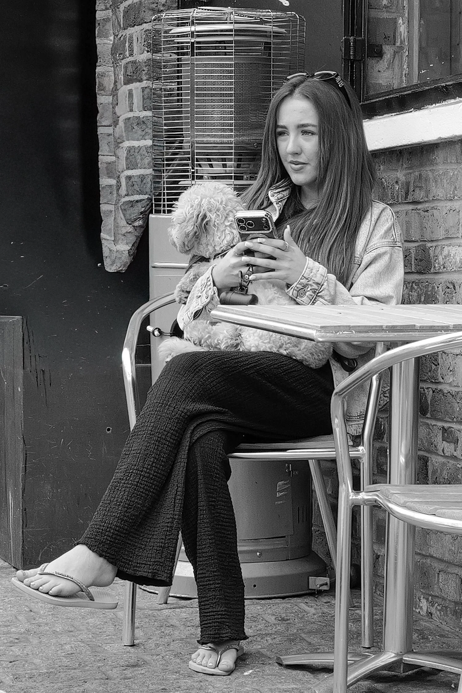 Une jeune femme assise à une table en extérieur, tenant un chien dans ses bras tout en regardant son téléphone. Elle porte des vêtements décontractés, des sandales et a des lunettes sur la tête. En arrière-plan, un mur en briques et une cage ou un ap