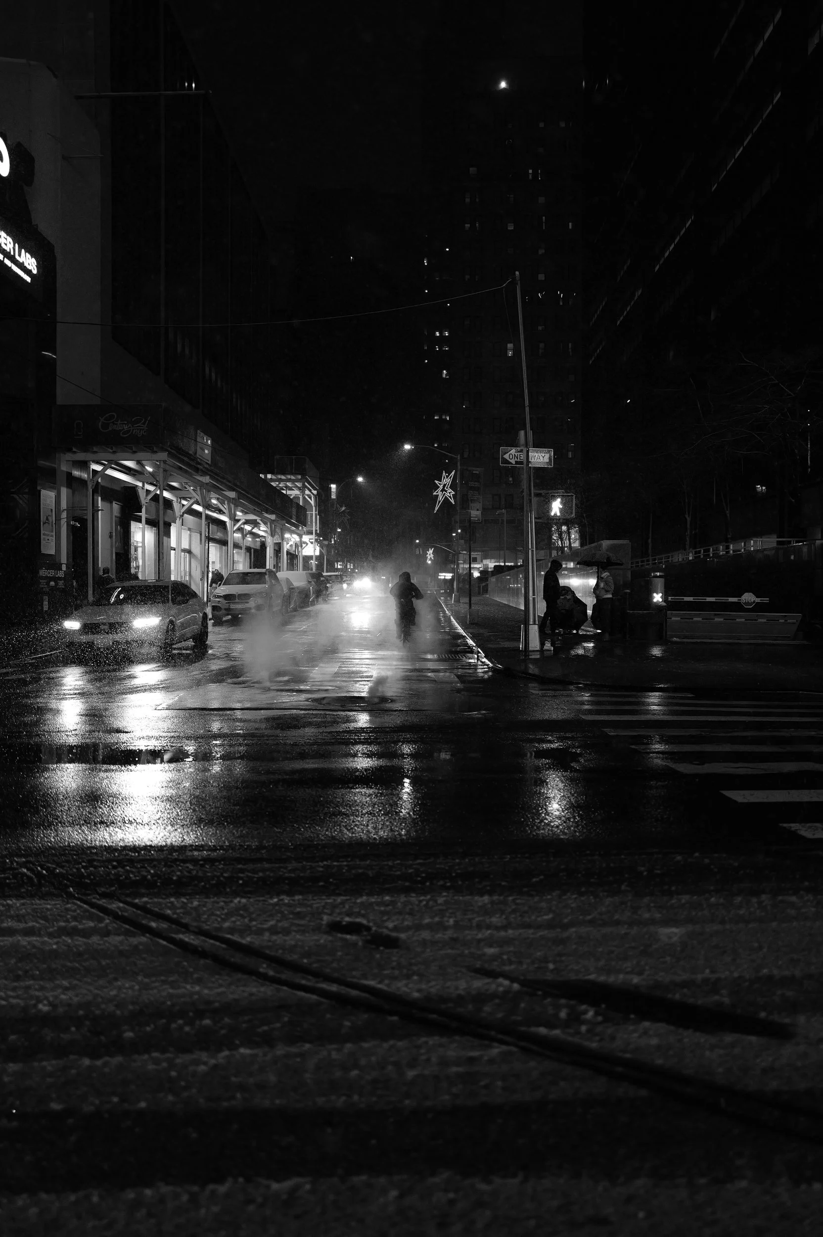 Une rue urbaine la nuit, sous la pluie, avec des personnes portant des parapluies et des voitures stationnées.