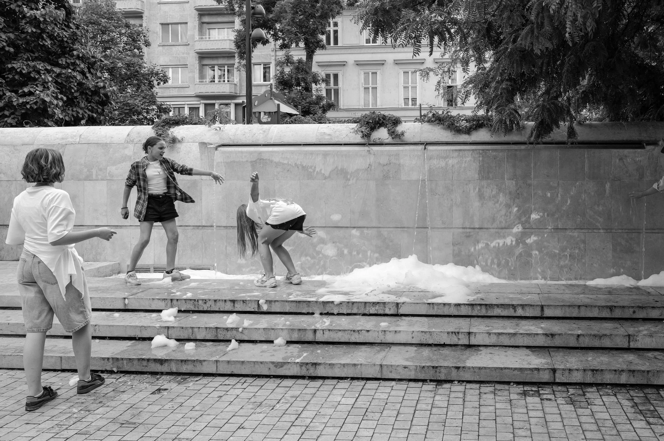 Quatre enfants jouent près d'une fontaine en pierre dans une zone urbaine avec des arbres et des bâtiments résidentiels en arrière-plan. Deux filles ont les cheveux longs, une porte une chemise à carreaux, une autre une veste blanche, un garçon a les