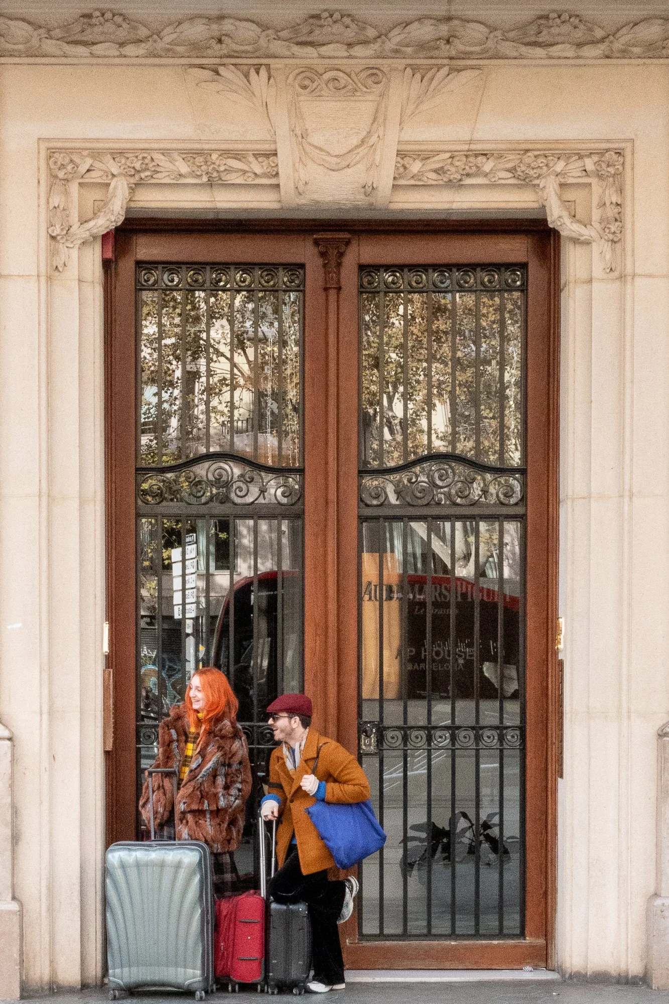 Deux personnes avec valises devant une porte en fer forgé et en bois, reflet de bâtiments et arbres, style architectural élégant.