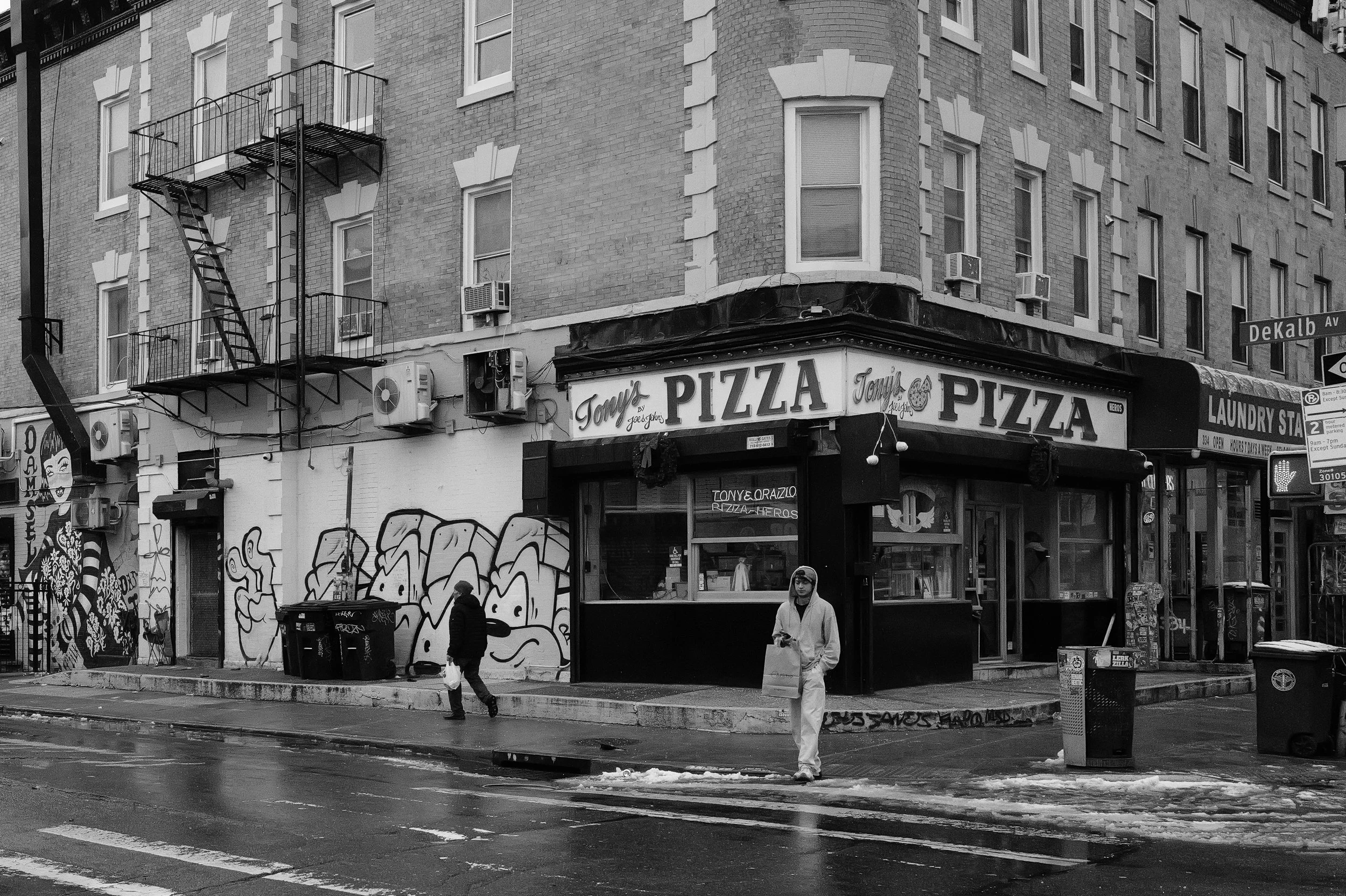 Vue d'une rue urbaine avec un restaurant de pizza nommé 'Tony's Pizza', des graffitis sur un mur, des personnes marchant sous la pluie, et un bâtiment à plusieurs étages avec des fenêtres et des unités de climatisation.