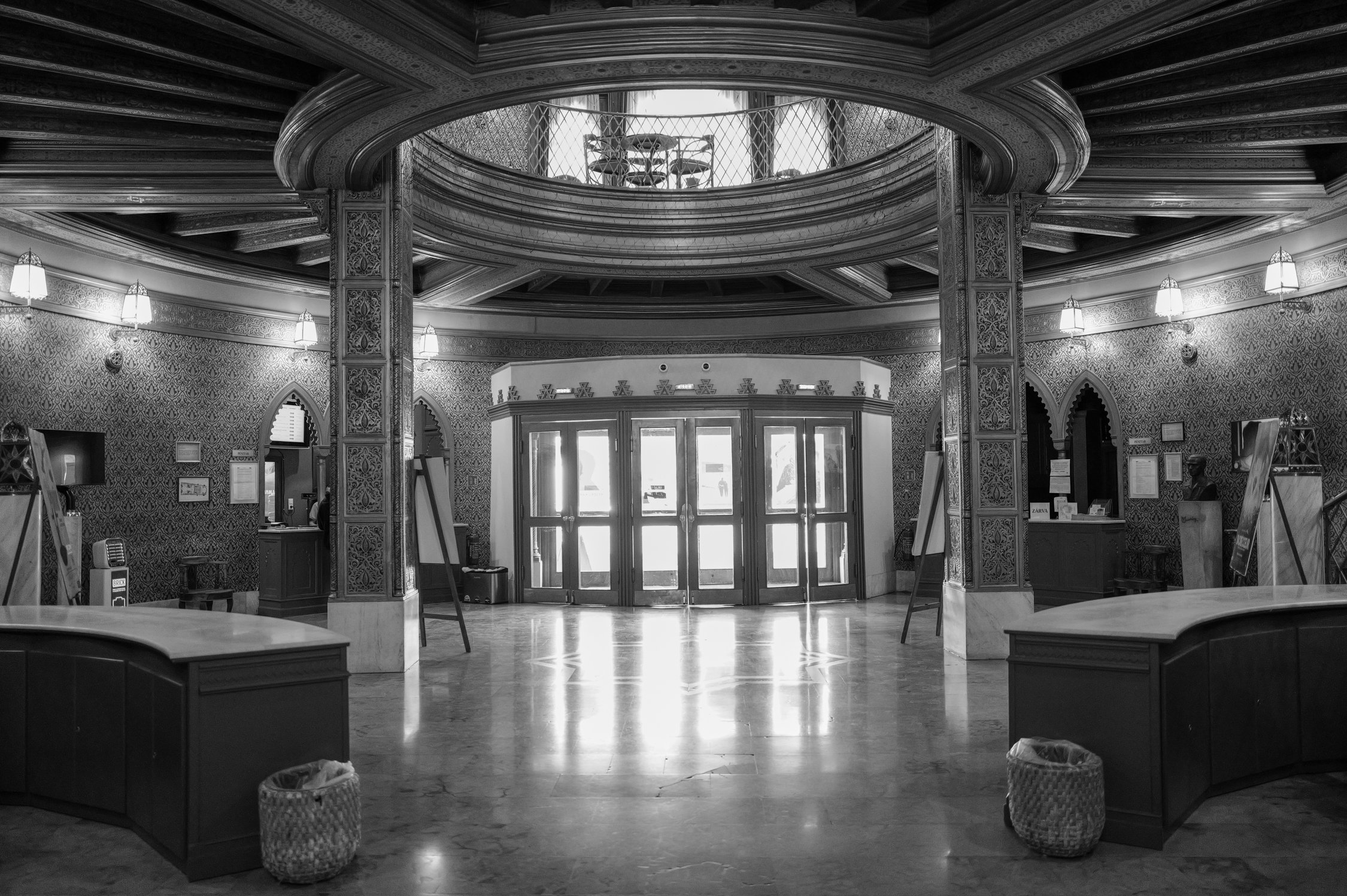 Une salle d'entrée ou un hall avec un design architectural sophistiqué, comprenant des murs ornés de motifs, des lampes suspendues, un plafond élaboré avec des moulures, et une porte d'entrée en bois avec de la lumière naturelle passant à travers. La