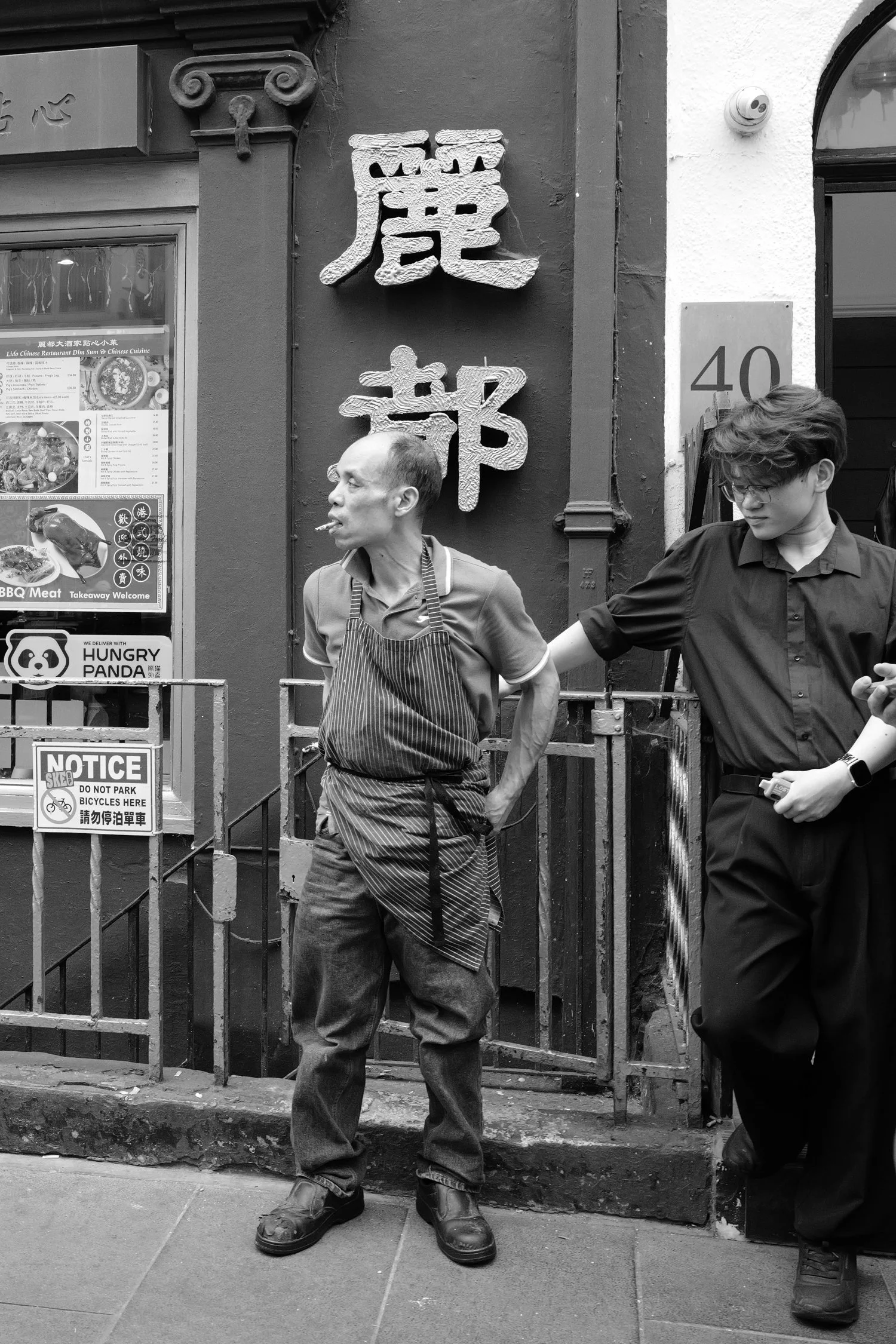 Deux hommes debout devant un restaurant chinois. L'un porte un tablier rayé, l'autre une chemise noire. Éléments de signalisation et enseigne en chinois sur le mur derrière eux.