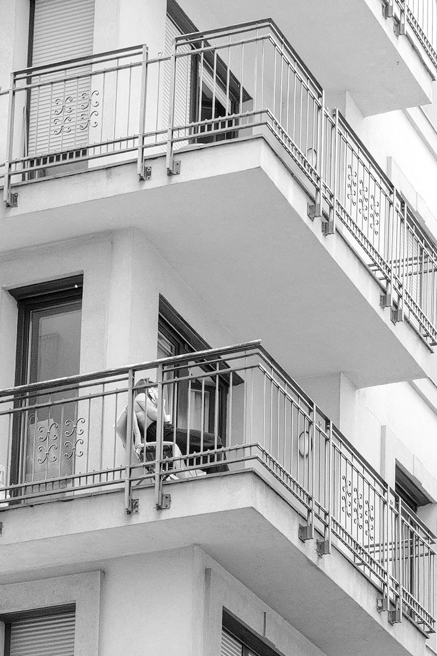 Vue en noir et blanc de balcons d'un immeuble résidentiel, avec une personne assise sur un fauteuil sur le balcon du milieu