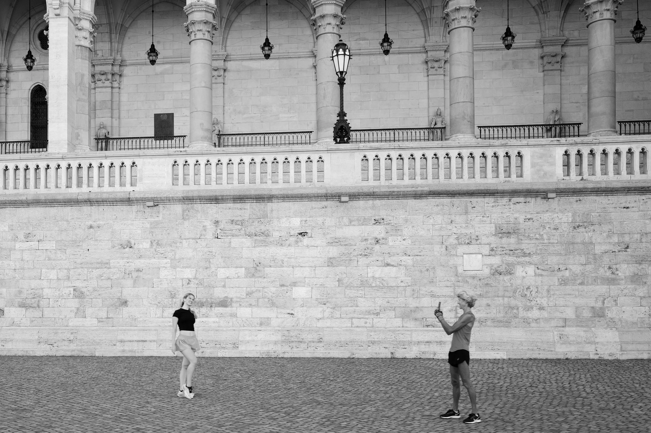 Deux femmes discutent et prennent une photo devant un mur en pierre, au centre d'une architecture classique avec des colonnes et des lampadaires.