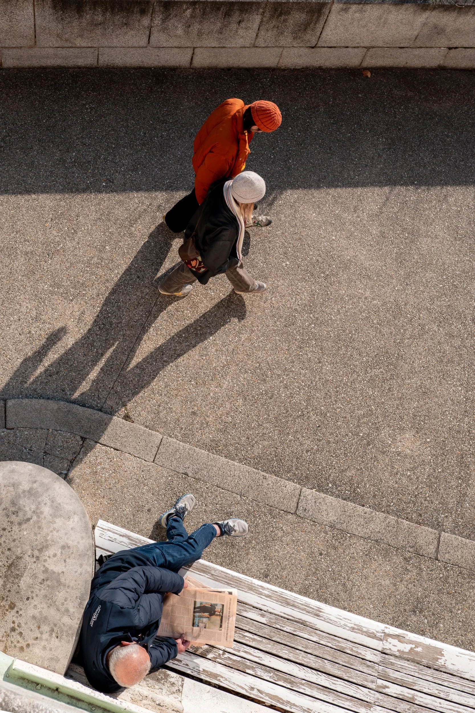 Une personne âgée lisant un journal assise sur un banc en bois, à côté d'une personne plus jeune. Deux autres personnes marchent sur le trottoir en direction opposée, dont une portant un manteau orange et un bonnet orange, et une autre portant un man