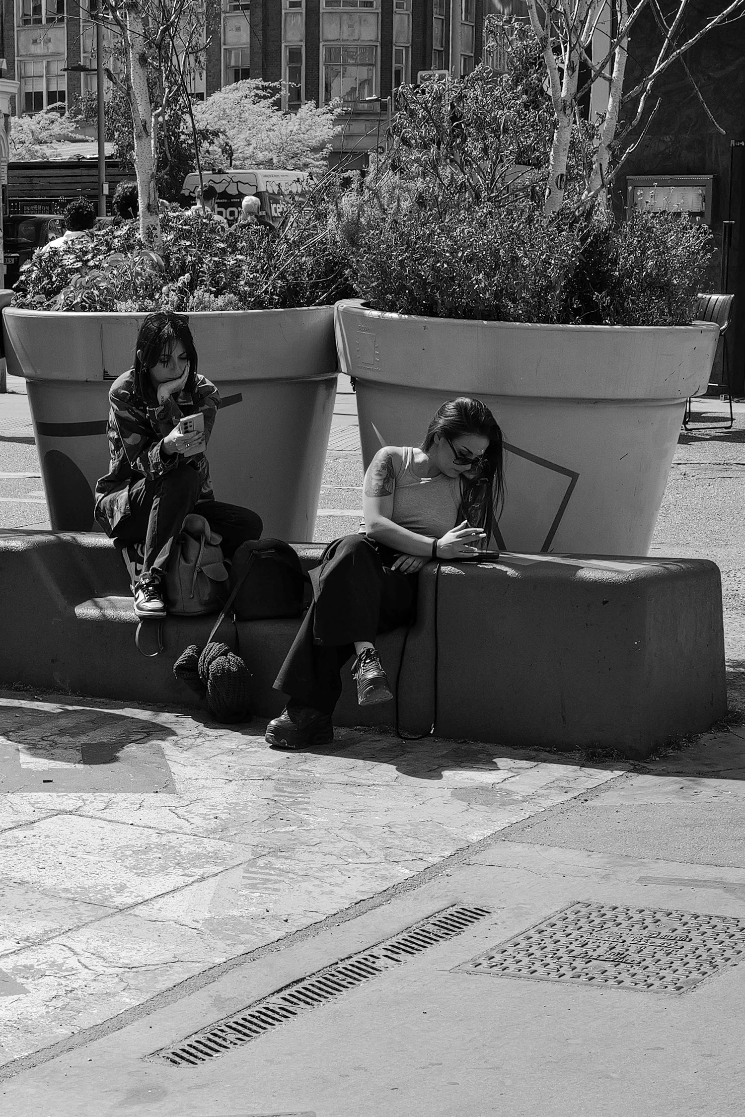 Deux femmes assises sur un banc en ville, utilisant leur téléphone portable. L'une porte un t-shirt à manches courtes et des lunettes, l'autre un pantalon camouflage et un sweat-shirt. Il y a deux grands pots avec des plantes derrière elles et une su