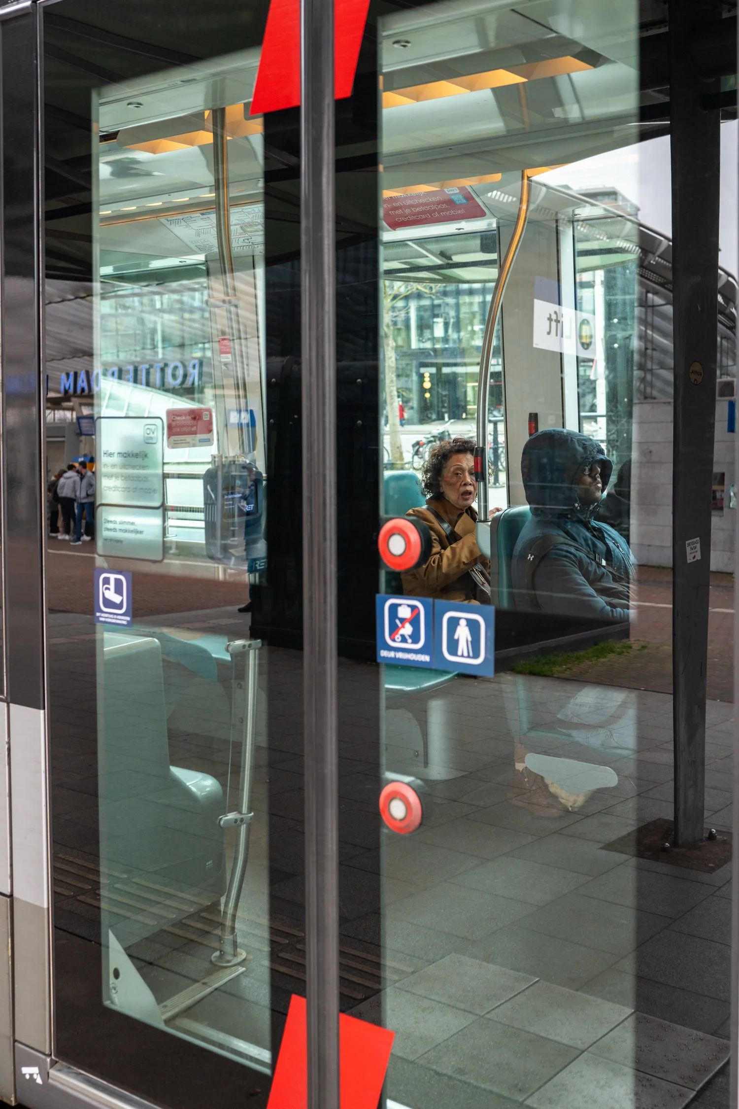 Une vue à travers une porte en verre montrant deux personnes assises à l'intérieur d'une station de bus ou de train, un homme portant une capuche et une femme avec des cheveux courts. À l'extérieur, il y a des arbres, des voitures, et des bâtiments.