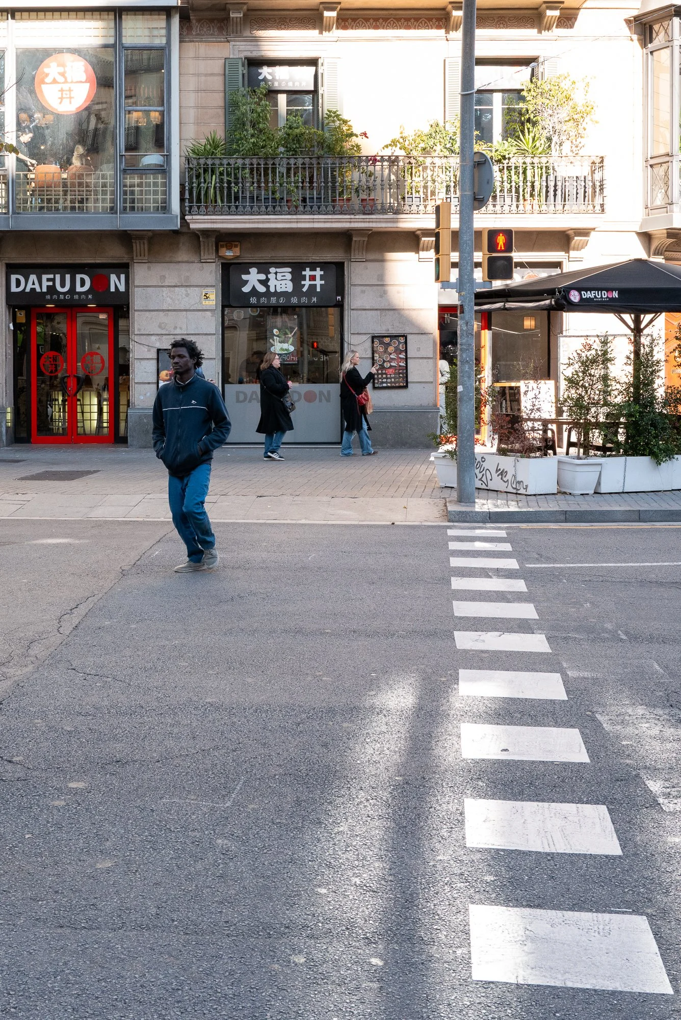 Une rue animée avec des personnes traversant un passage piéton devant un restaurant japonais appelé DAFUDON, avec des plantes en terrasse et un bâtiment en pierre avec des fenêtres.